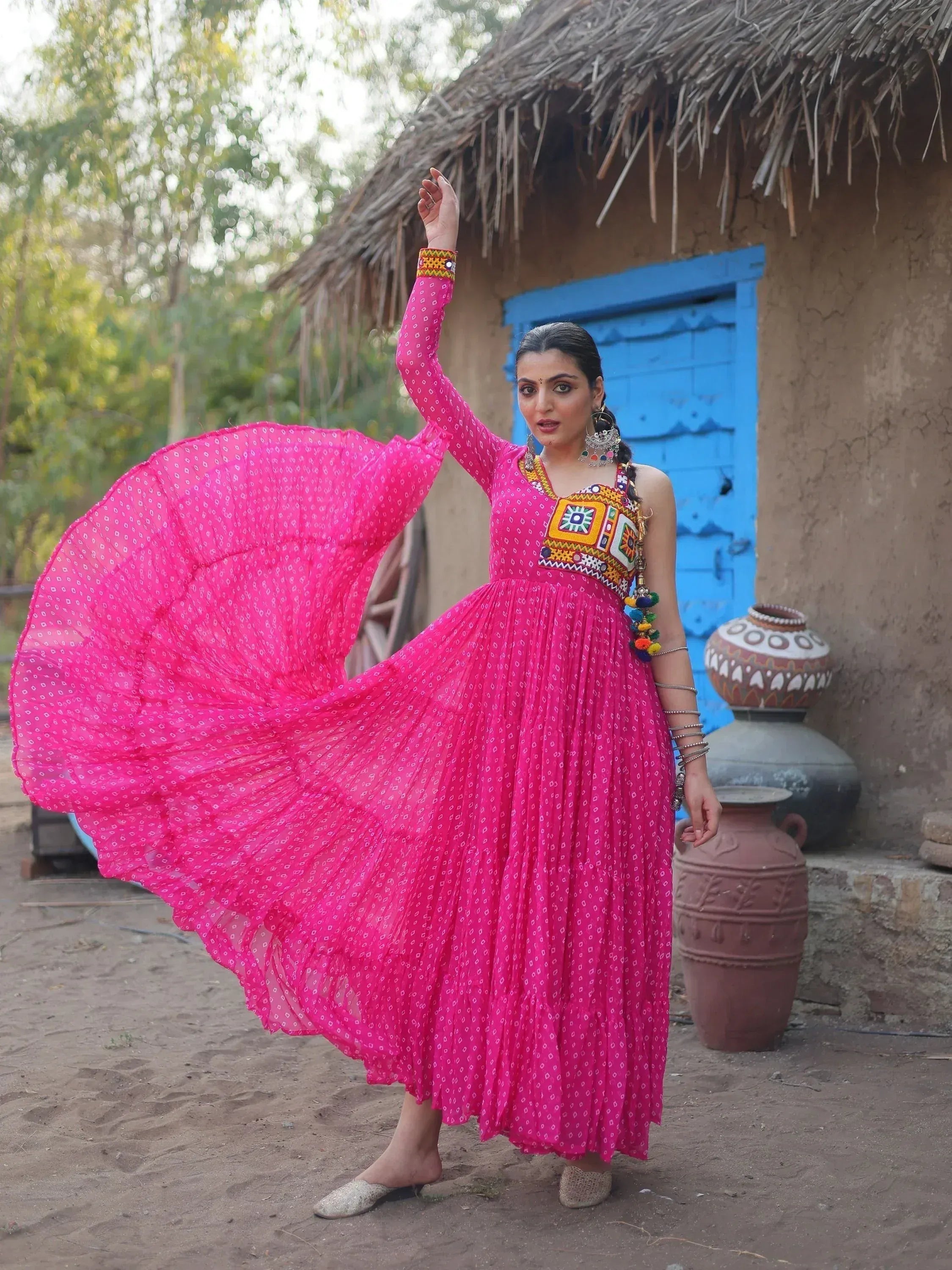Vibrant pink ethnic gown with mirror work, worn by woman dancing near rustic hut, perfect for Indian festive occasions
