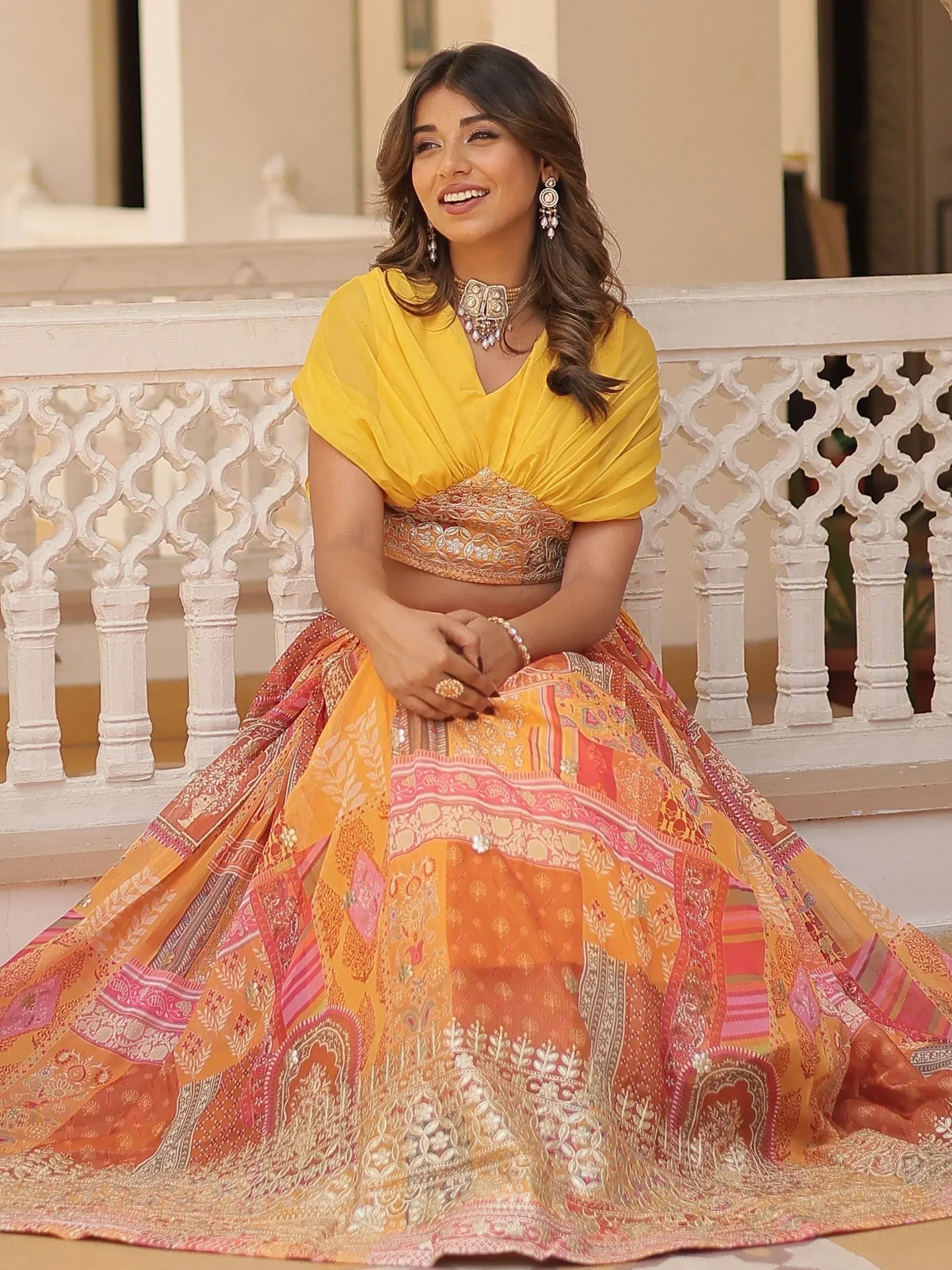 Woman in vibrant yellow and orange printed lehenga choli with embroidery, sitting on a balcony