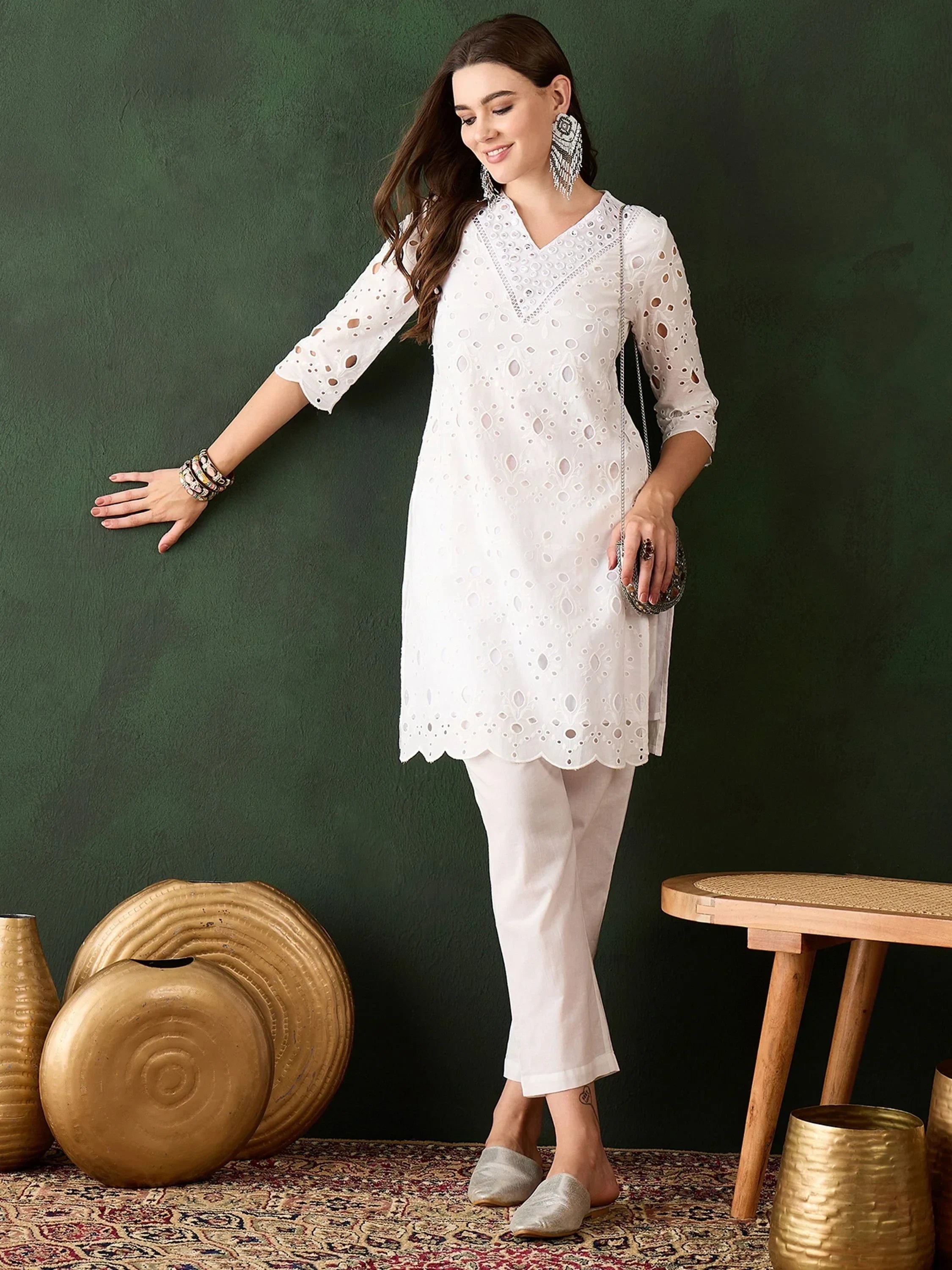 Woman in white embroidered kurti and pants, wearing ethnic earrings, standing on patterned rug