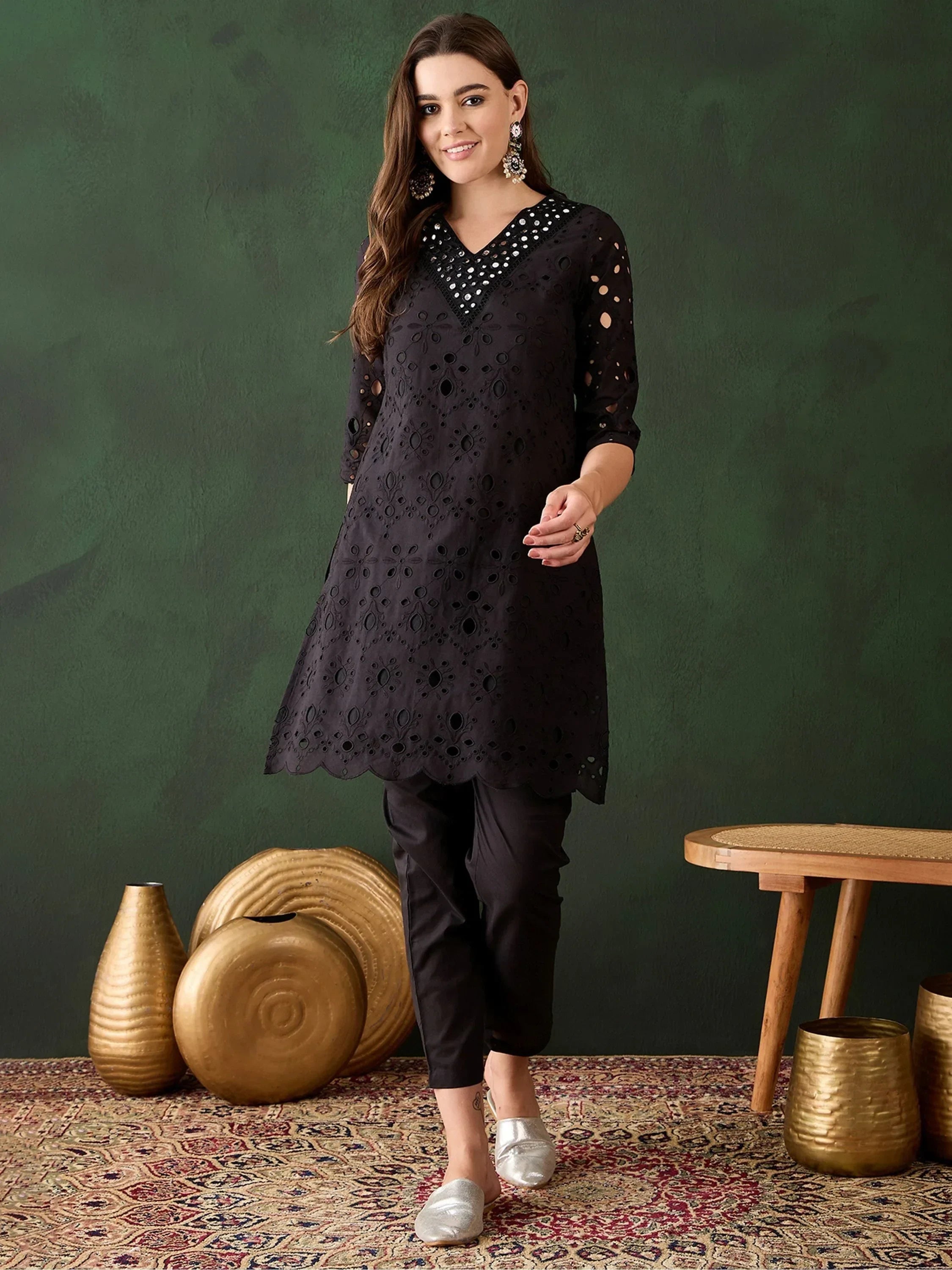 Woman wearing black embroidered kurti with pants, silver shoes, and ethnic earrings, standing indoors