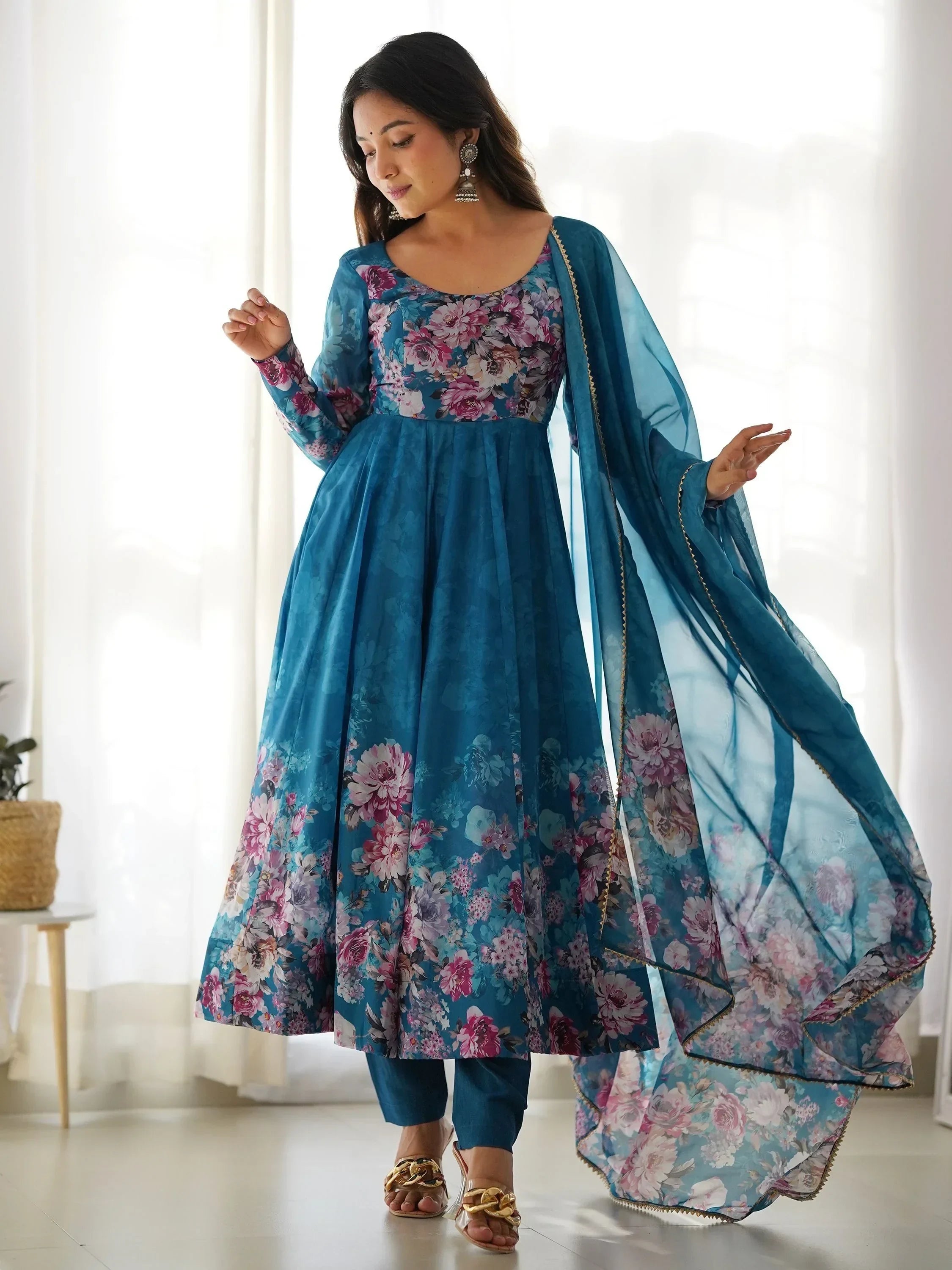 Woman in blue floral Anarkali suit with sheer dupatta and gold sandals, posing indoors