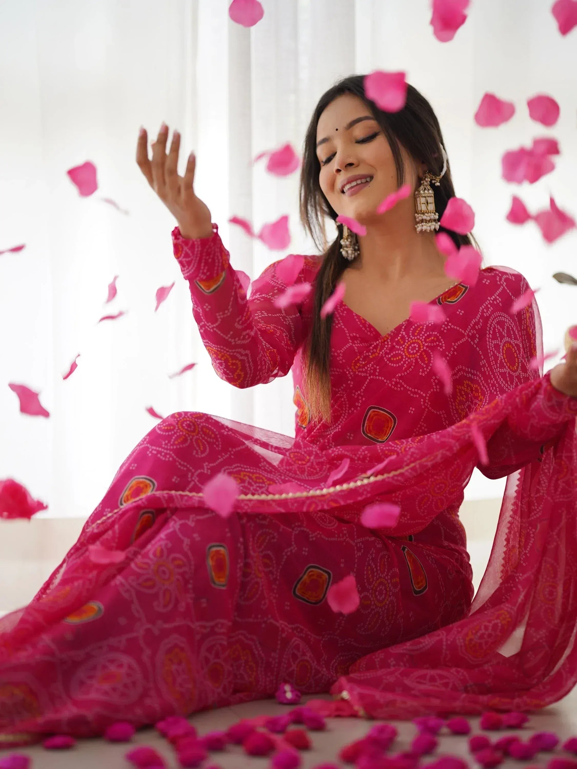 Woman in vibrant pink ethnic gown with floral print, surrounded by falling rose petals, styled with jhumka earrings.