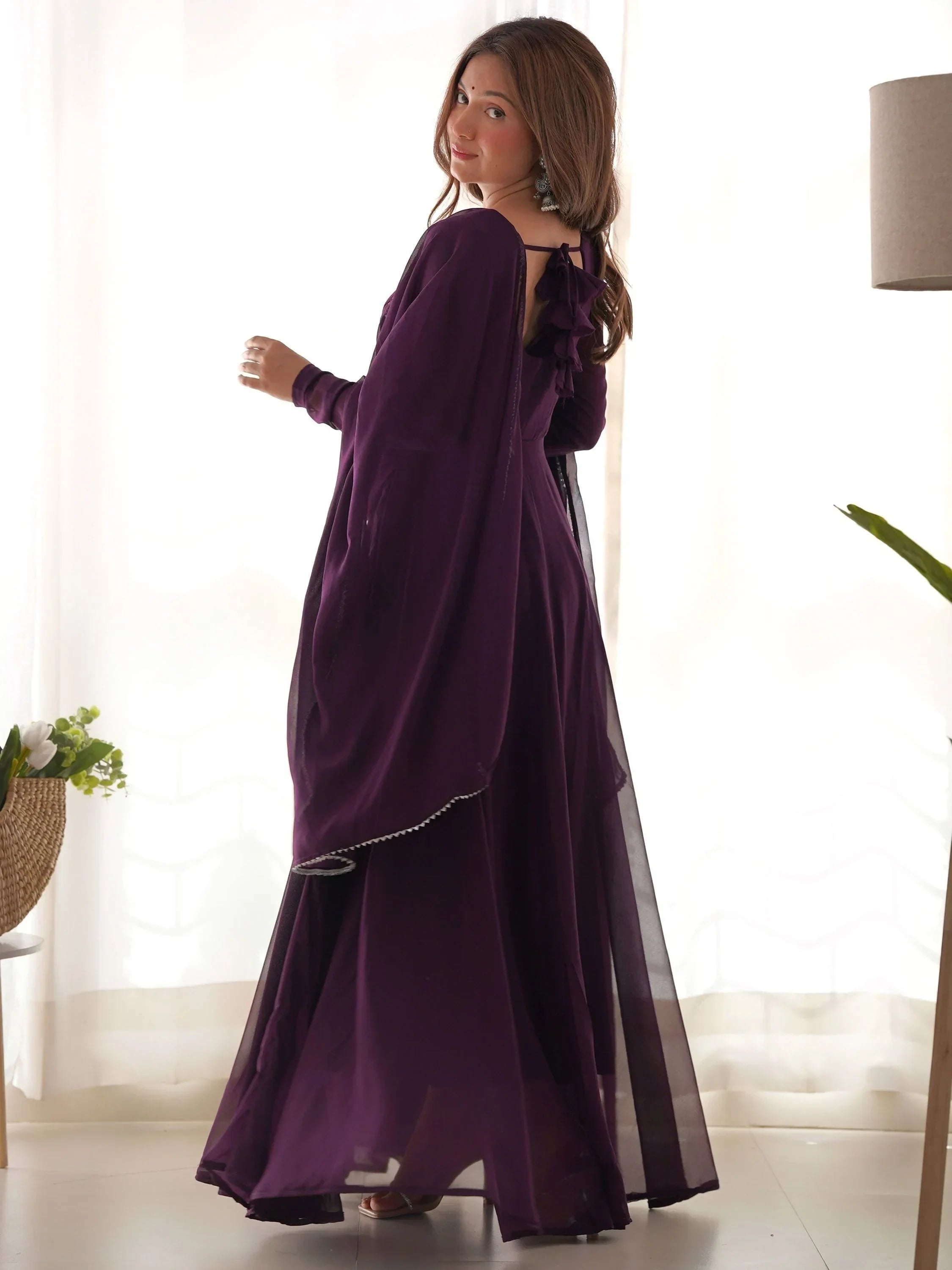 Woman in a deep purple ethnic gown with ruffle back and dupatta, standing indoors