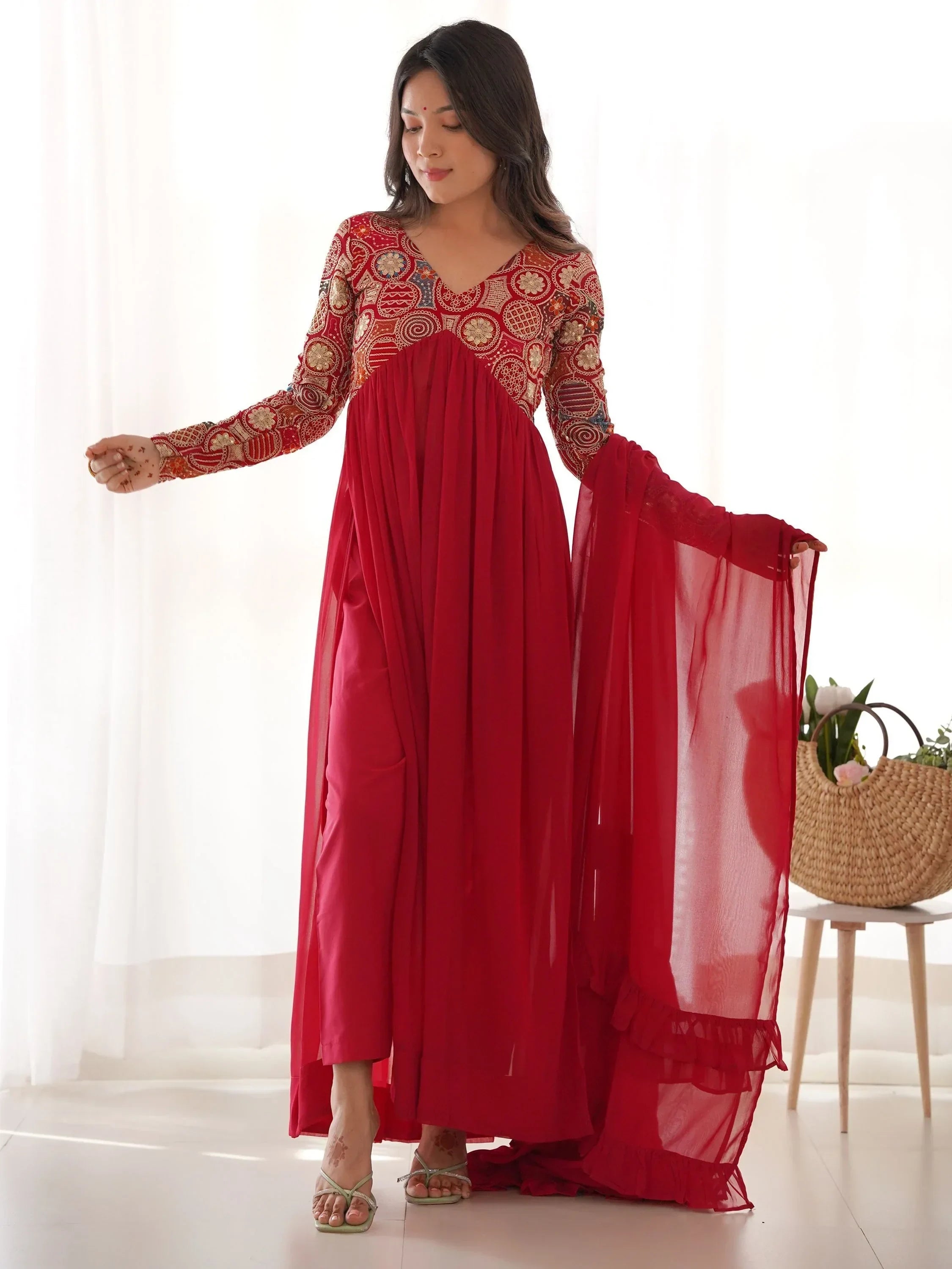 Woman in red embroidered ethnic gown with sheer dupatta, standing indoors