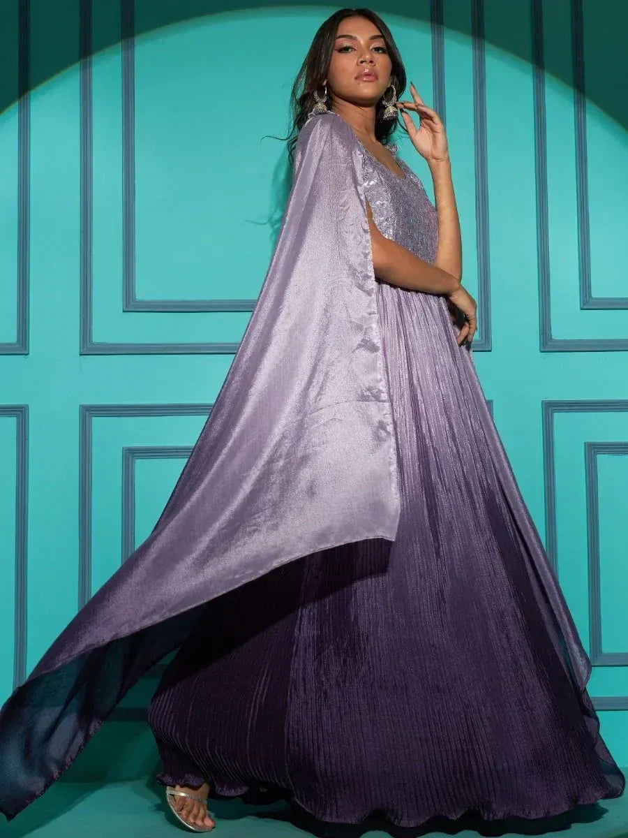 Woman in a purple pleated ethnic gown with cape sleeves, standing against teal wall