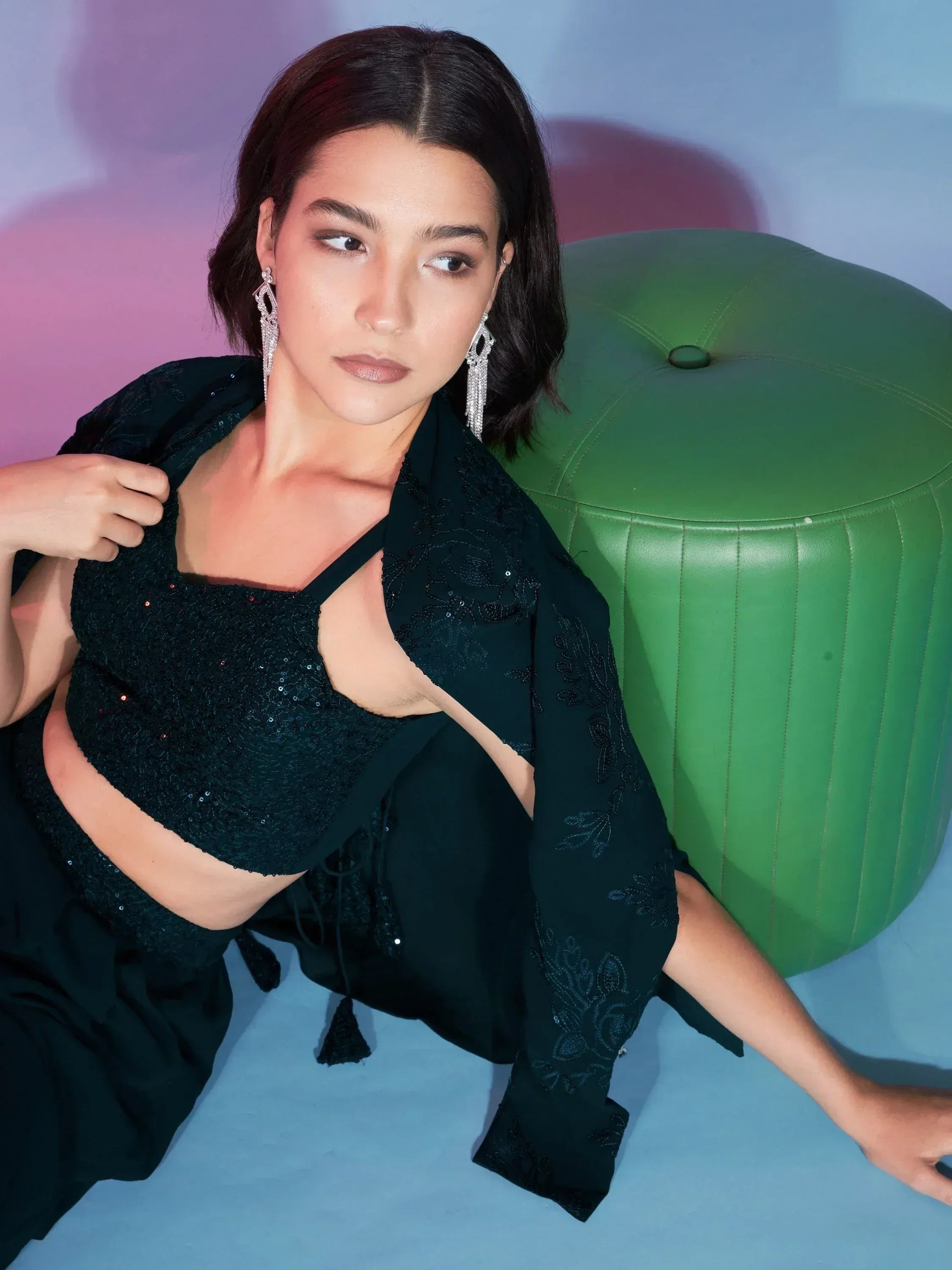 Woman in black sequin ethnic crop top with embroidered jacket, stylish earrings, seated on green ottoman