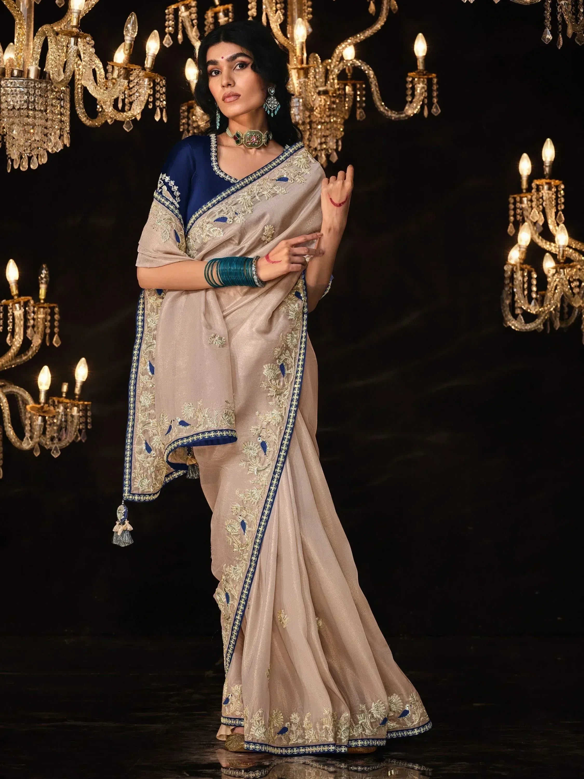 Elegant beige saree with blue and gold embroidery, paired with matching blouse, showcased under chandeliers.