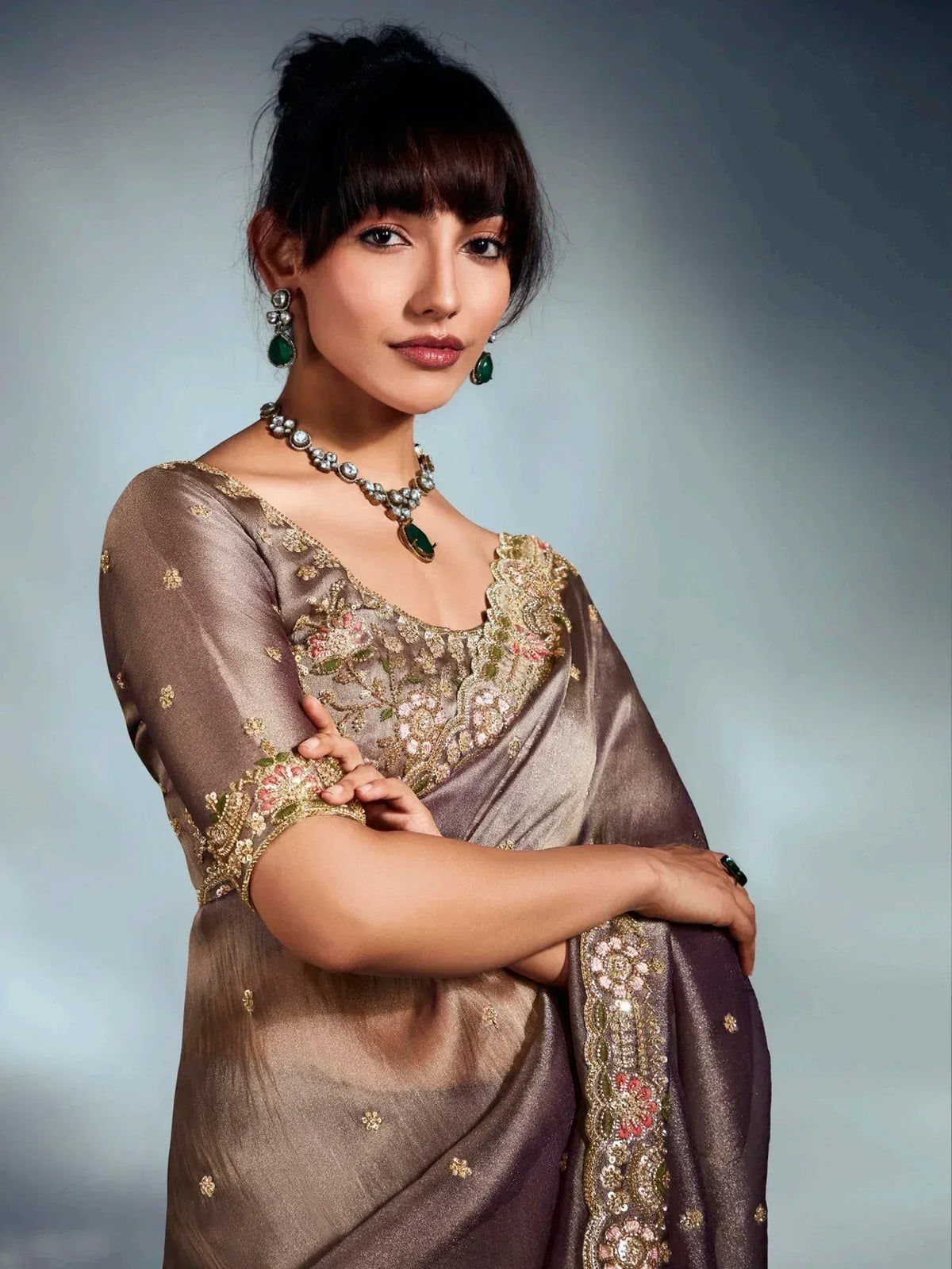 Elegant woman in embroidered brown saree with floral gold detailing, paired with green gemstone jewelry