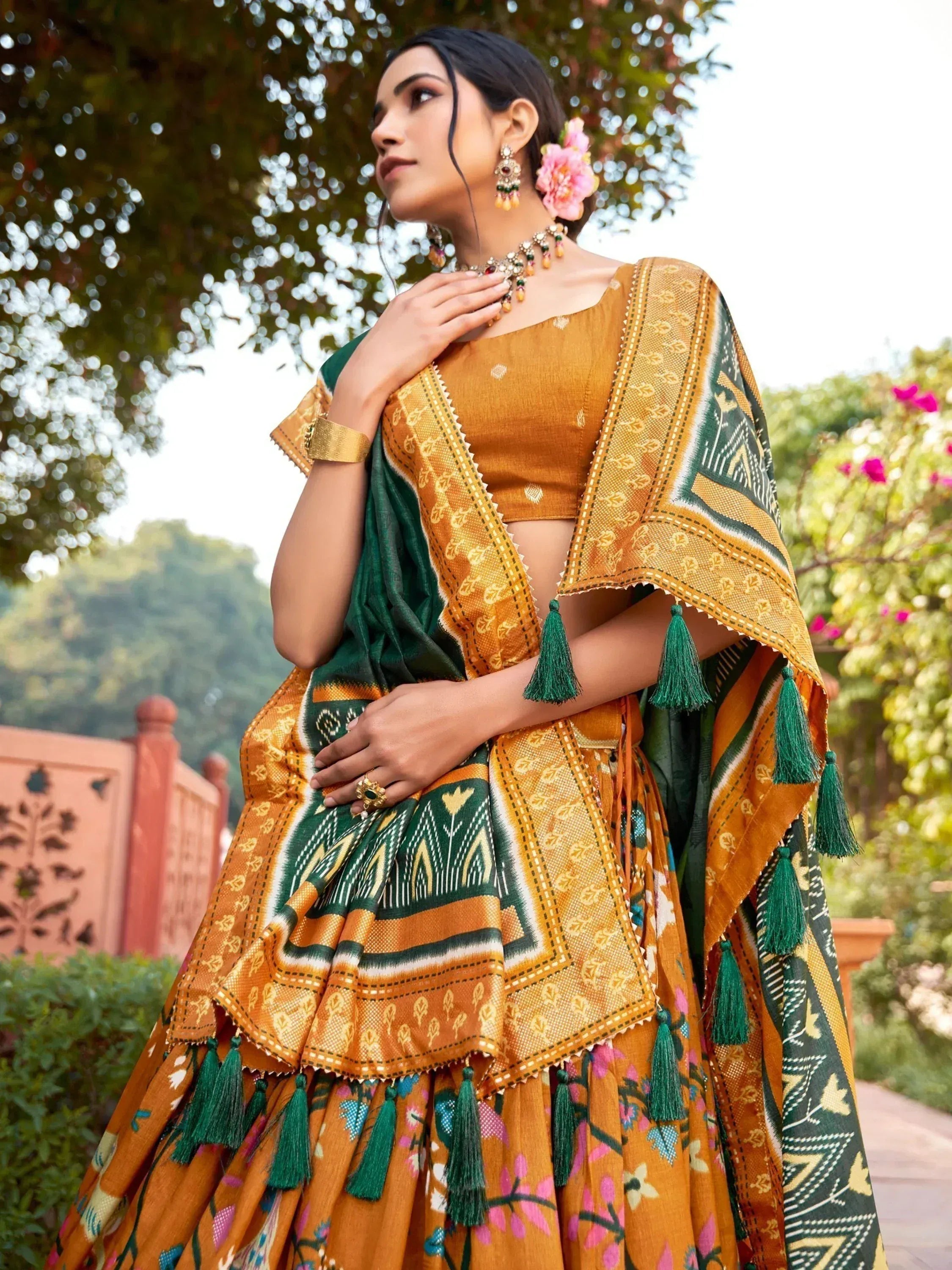 Woman in mustard and green ethnic lehenga choli with tassels, floral print, and traditional jewelry outdoors