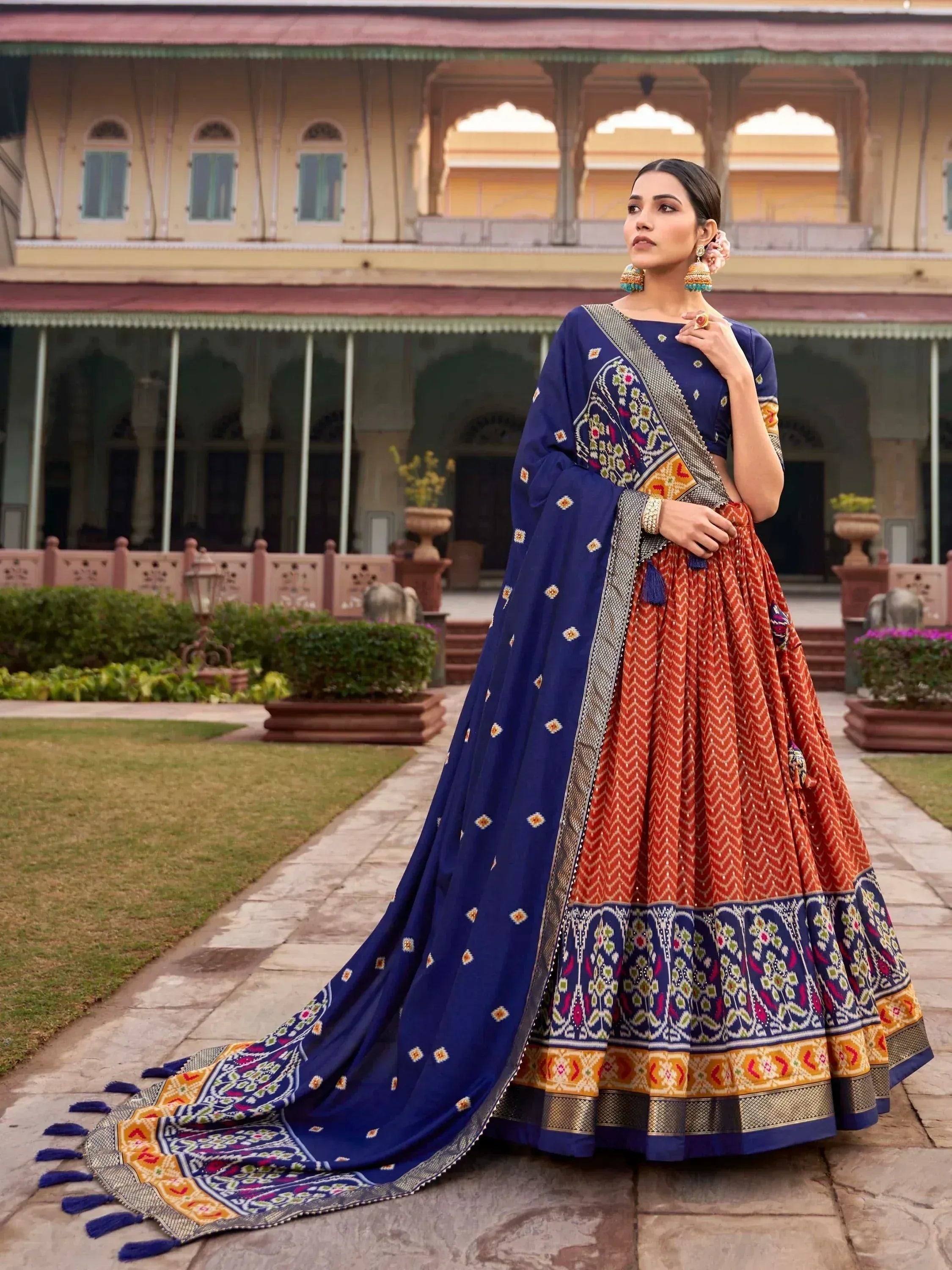 Woman in vibrant blue and orange ethnic lehenga choli with intricate embroidery, standing in palace courtyard