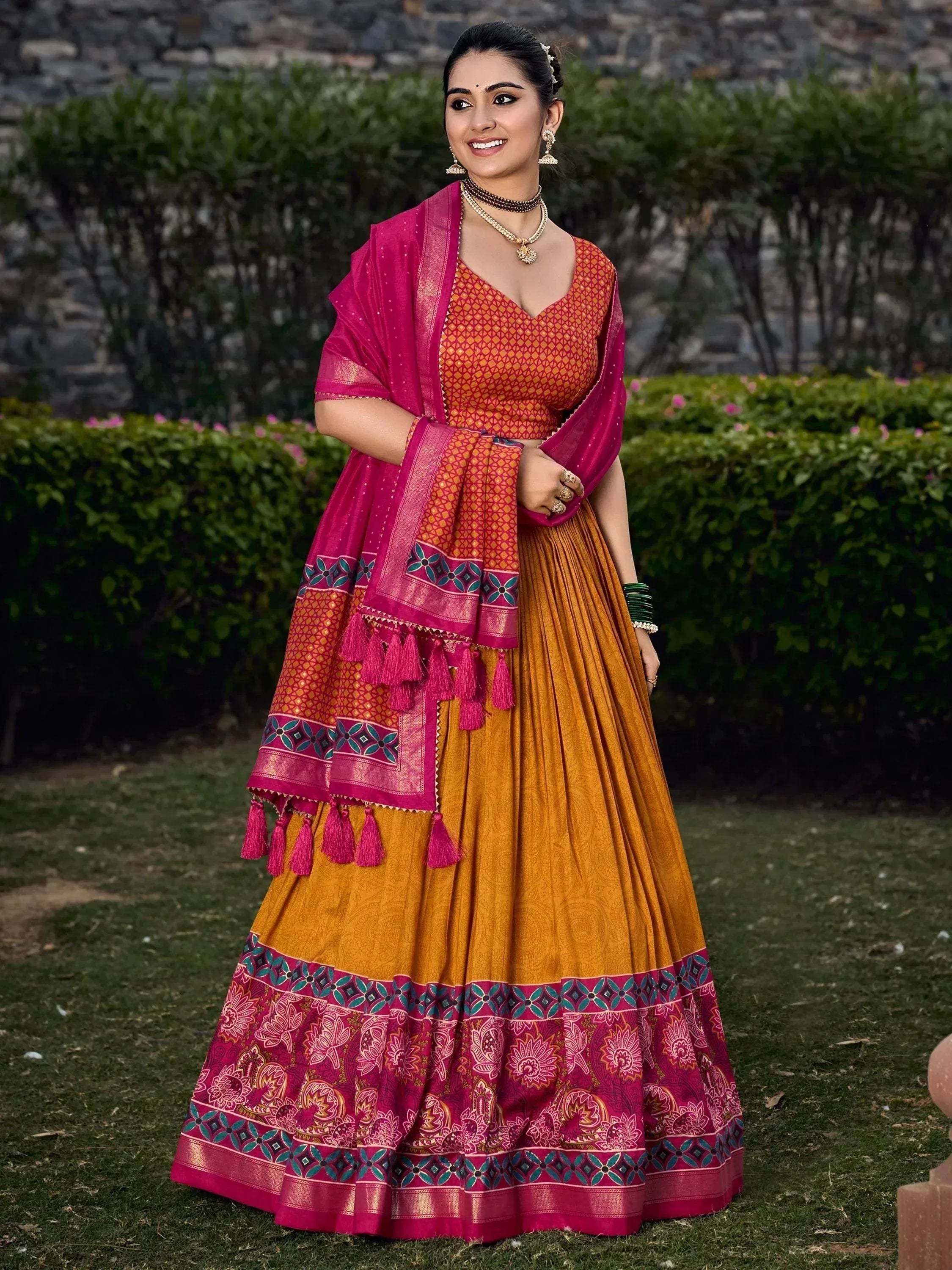 Woman in vibrant Indian lehenga choli with pink dupatta, floral border, and tassels, perfect for ethnic occasions.