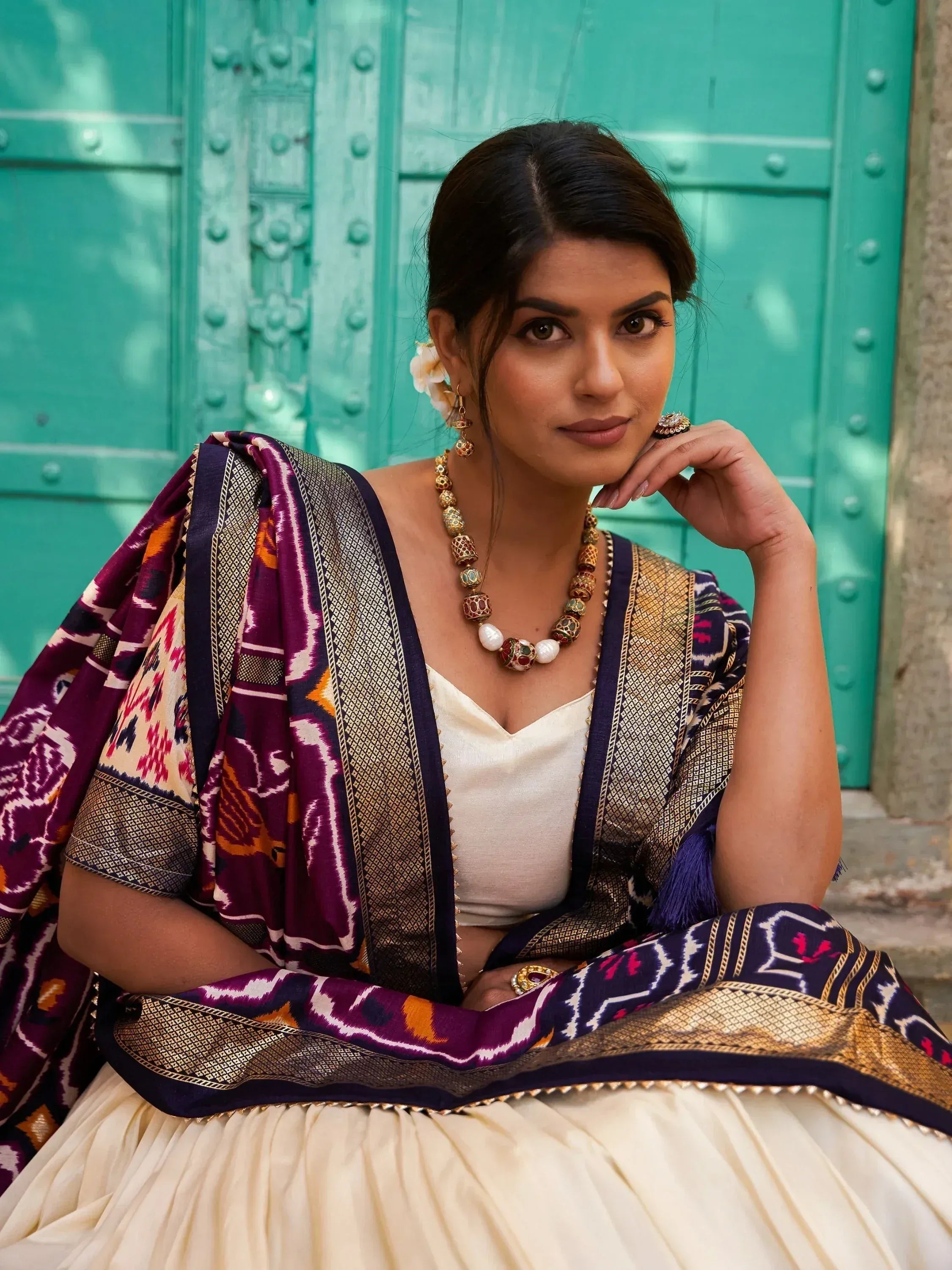 Woman in purple ikat lehenga choli with gold jewelry, posing against teal door