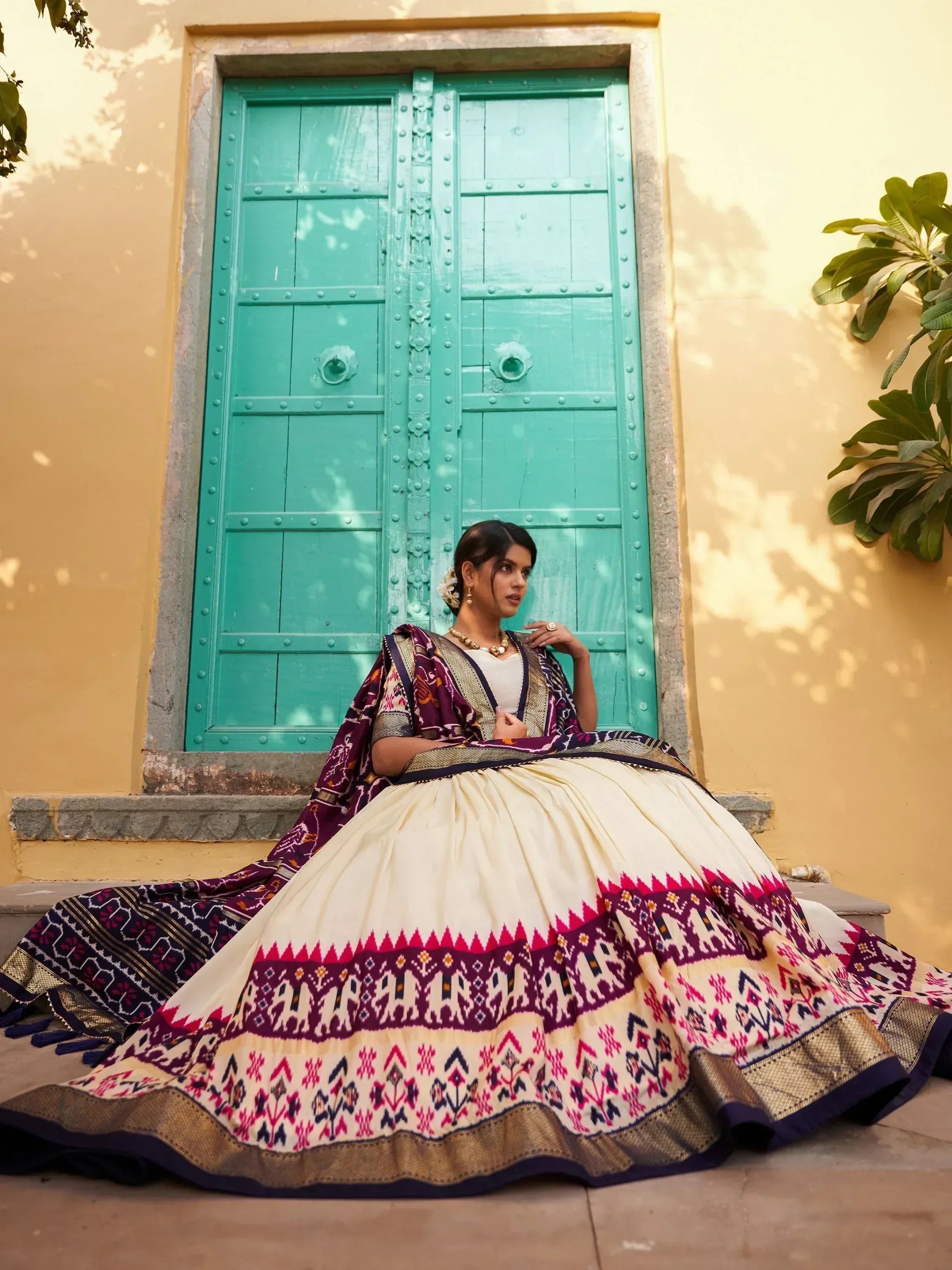 Woman in ethnic lehenga choli with vibrant ikat print, seated before turquoise door