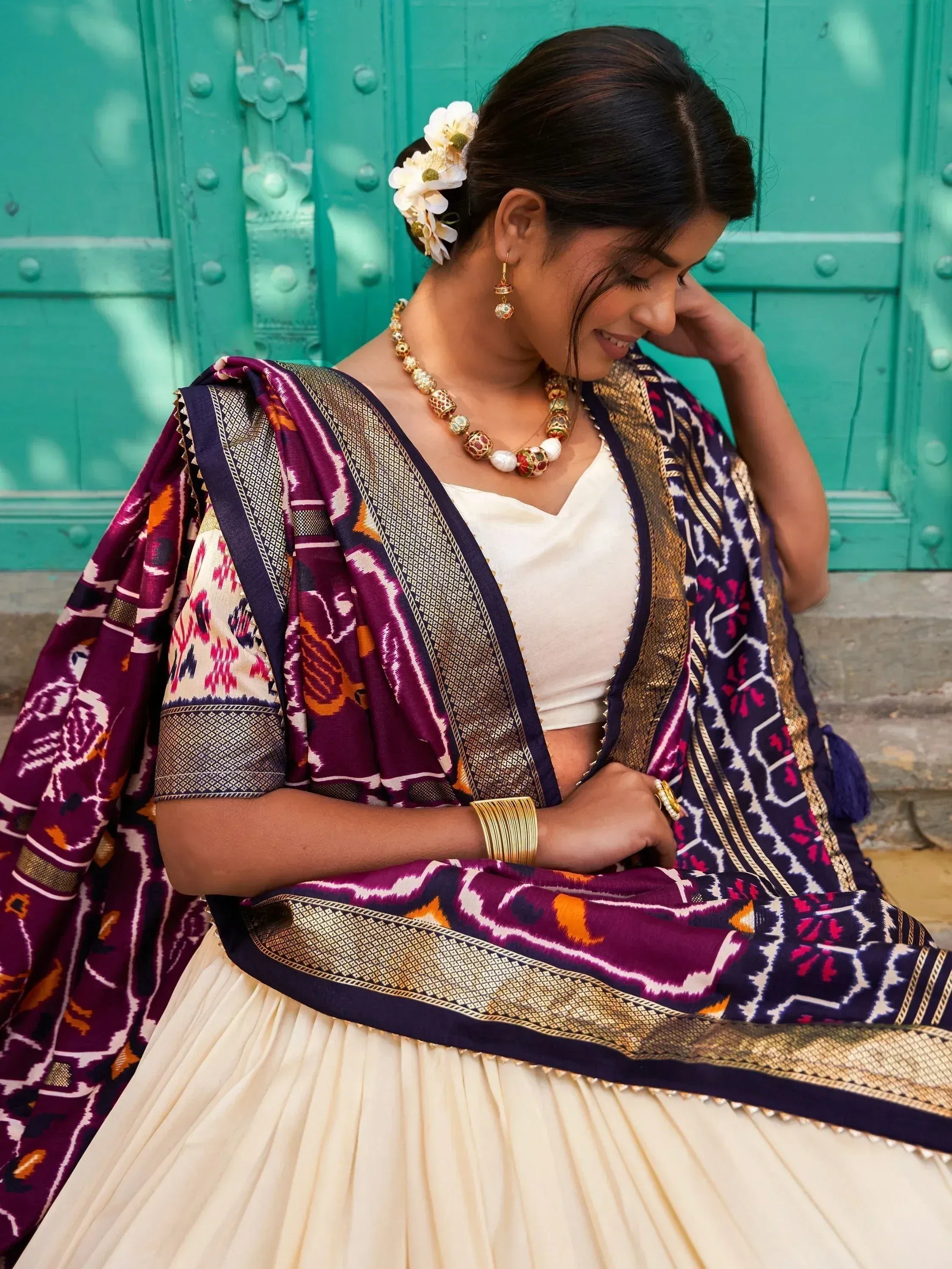 Woman in purple ikat saree with gold border, white blouse, floral hair accessory, and traditional jewelry against teal door