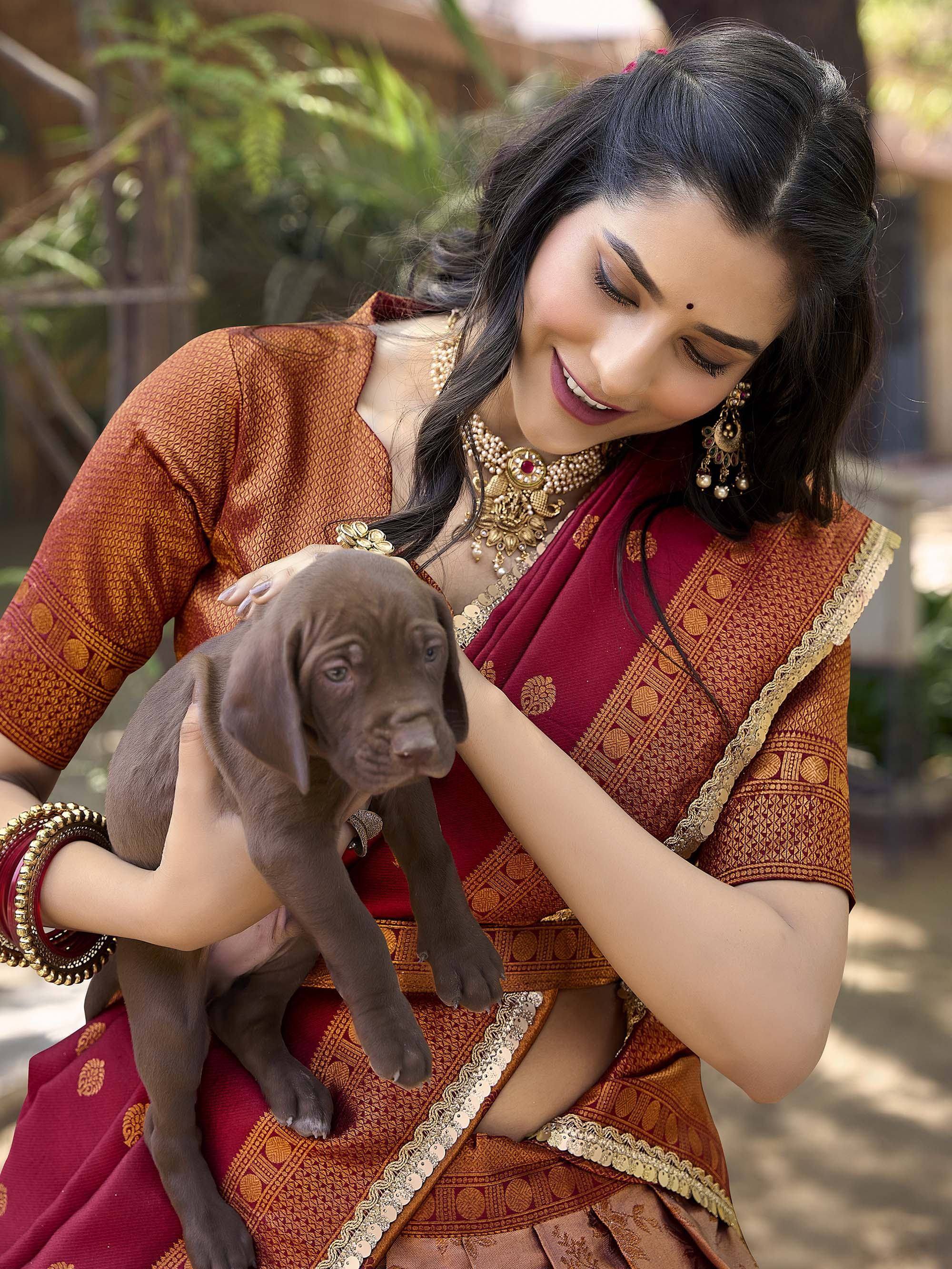 Woman in maroon and gold ethnic saree holding a puppy, traditional Indian attire with jewelry