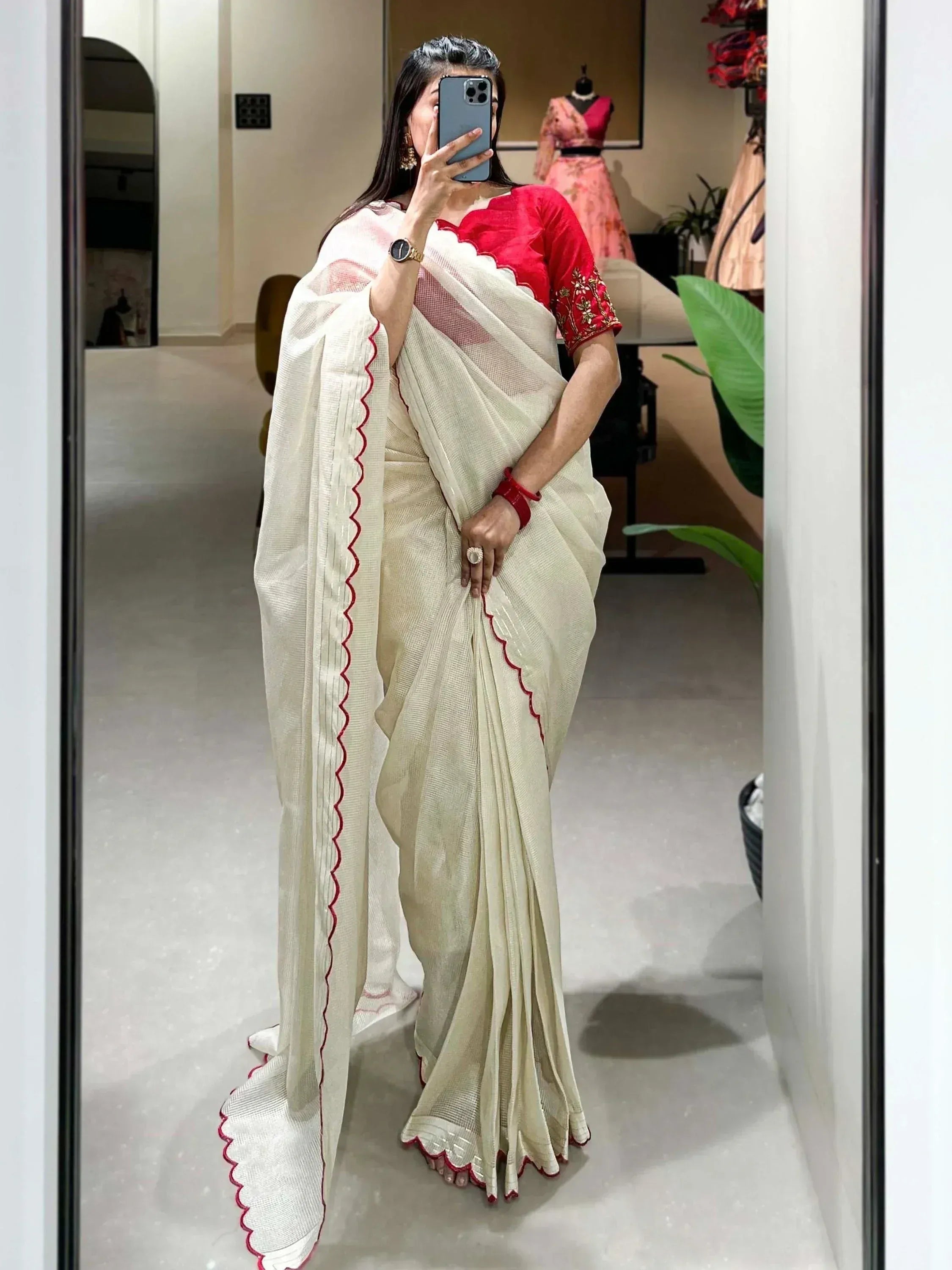 Woman in off-white saree with red scalloped border and matching red blouse, taking mirror selfie in boutique