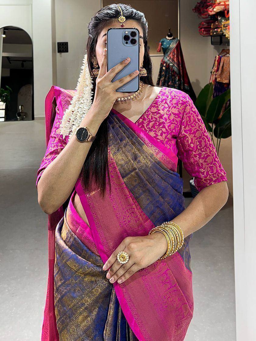Woman in vibrant pink and blue designer saree with gold embroidery, traditional jewelry, and floral hair accessory.