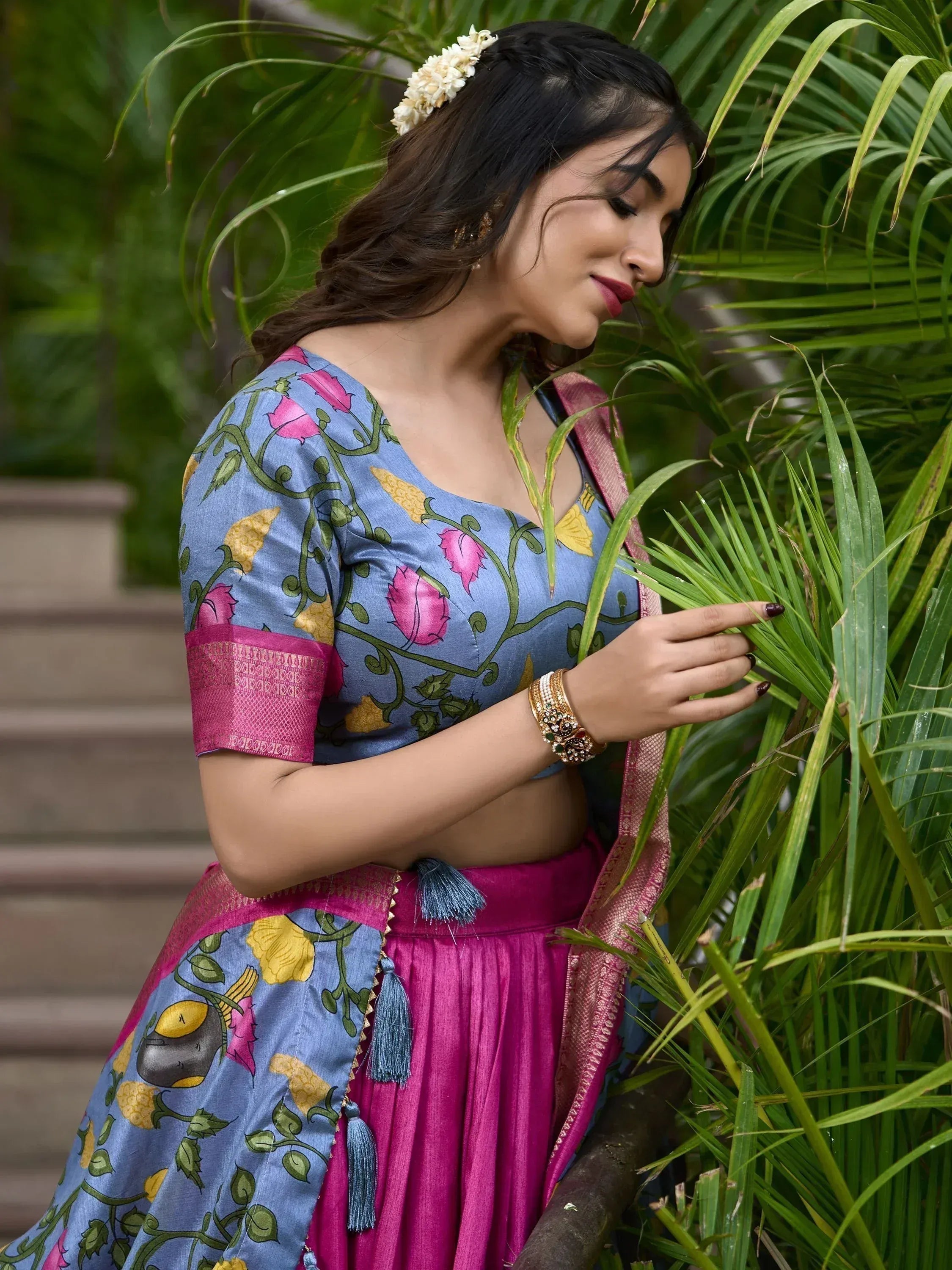 Woman in blue floral lehenga choli with pink skirt, gold jewelry, and flower hair accessory, standing near green palm leaves.
