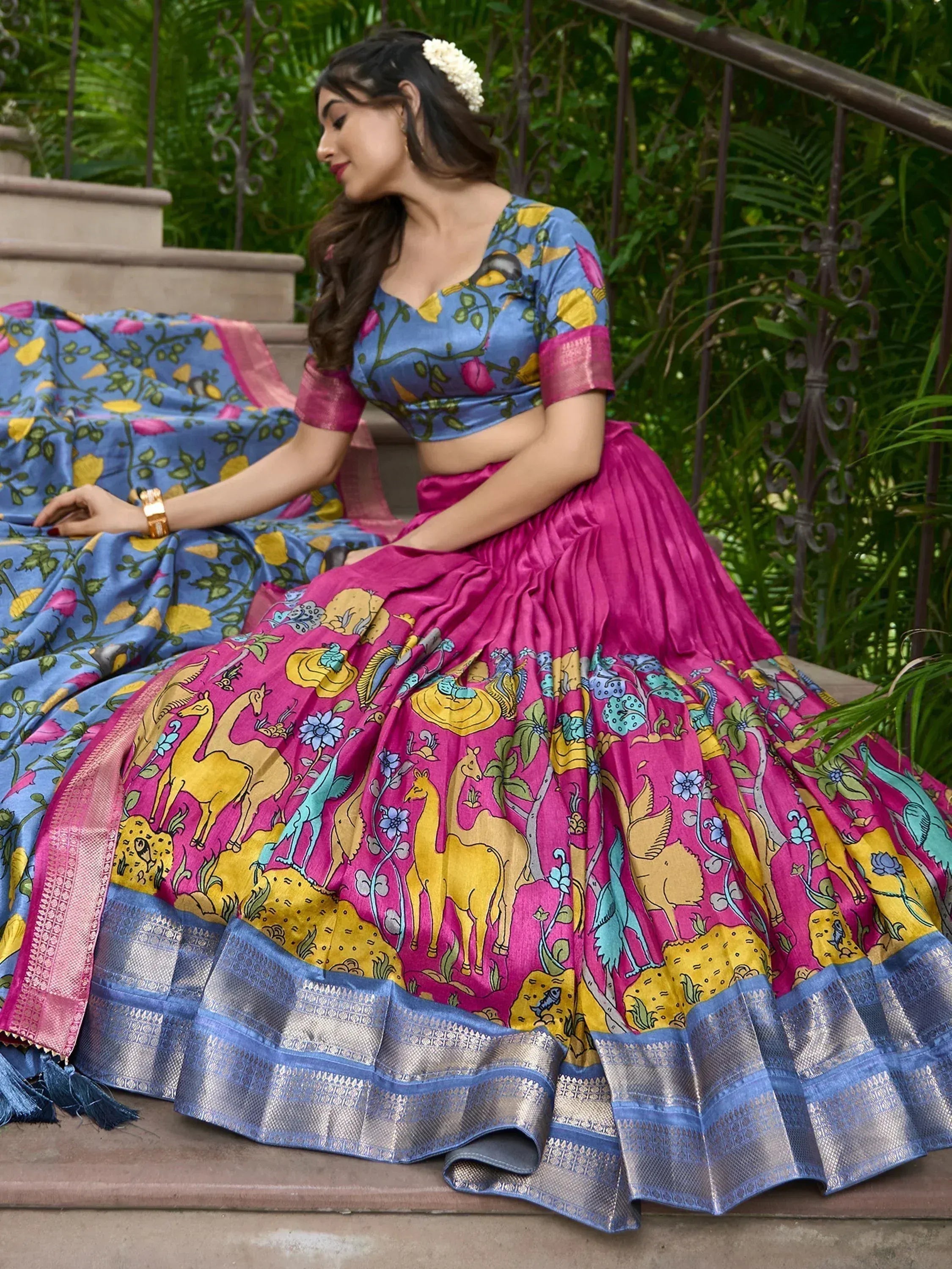 Woman in vibrant pink and blue printed lehenga choli with floral and animal motifs, sitting on garden steps