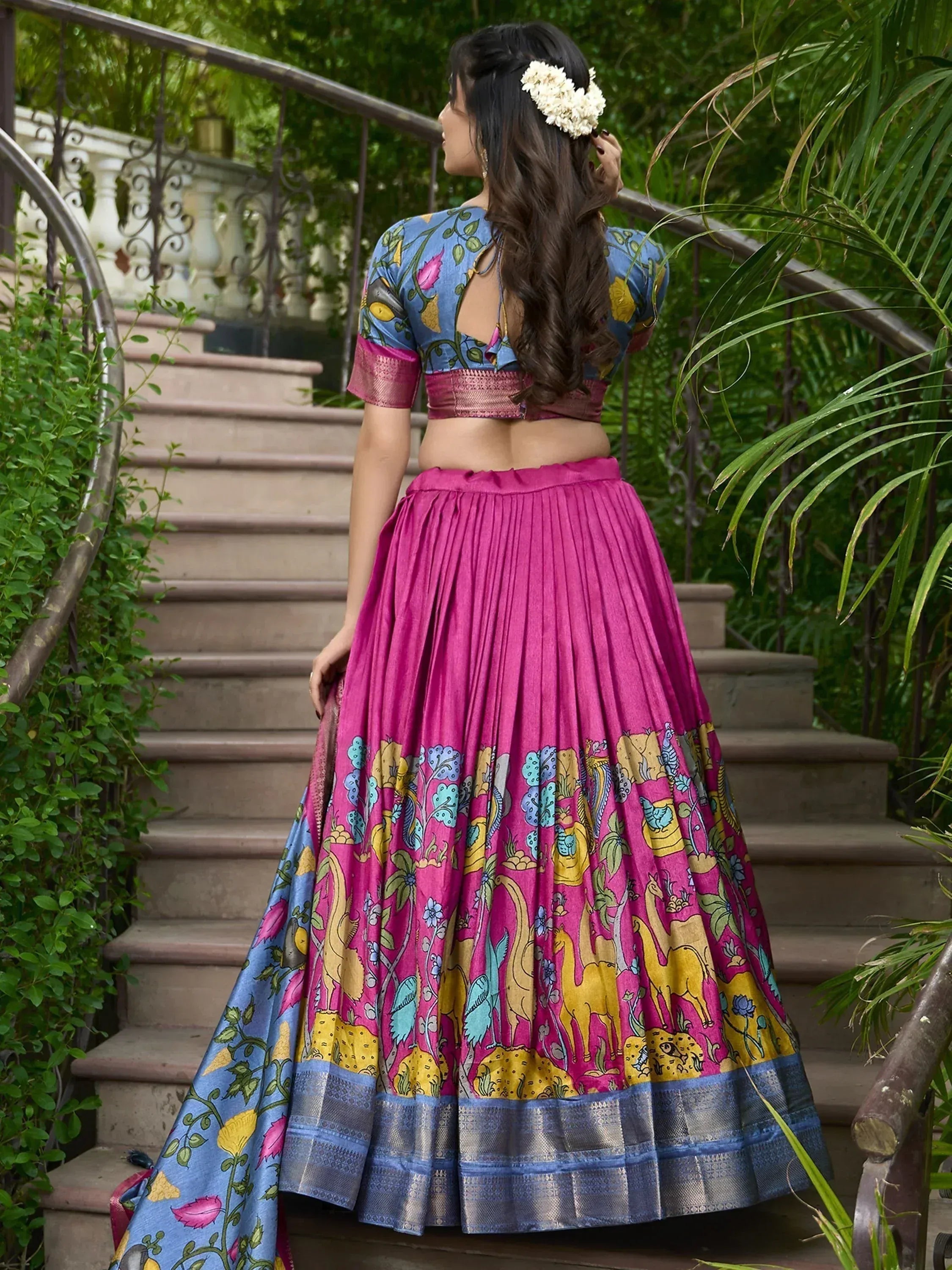 Woman in vibrant pink and blue printed lehenga choli with floral and animal motifs, standing on garden stairs