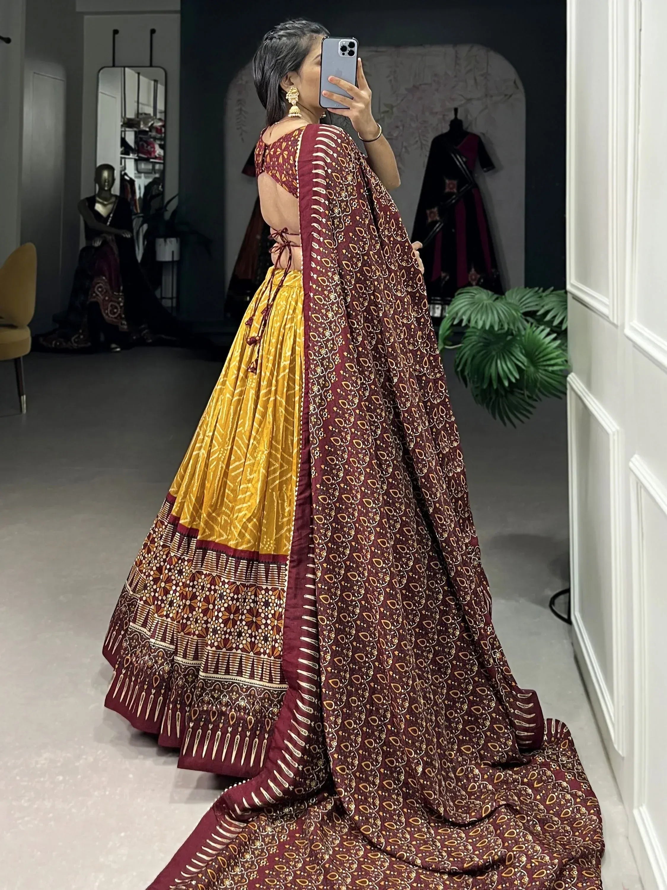 Woman in maroon and mustard printed lehenga choli with backless blouse, taking mirror selfie in boutique