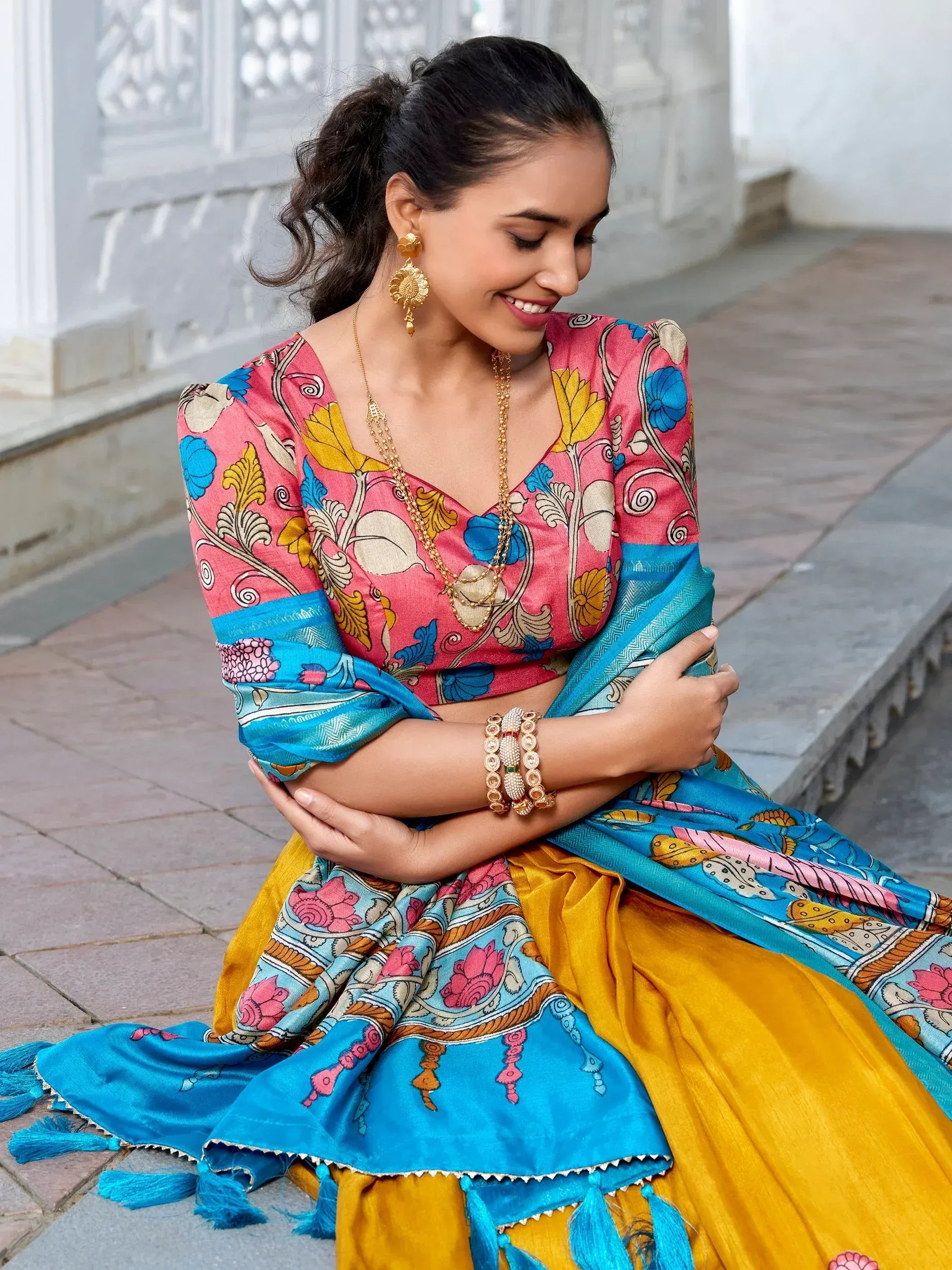 Woman in vibrant floral lehenga choli with blue dupatta, gold jewelry, sitting outdoors
