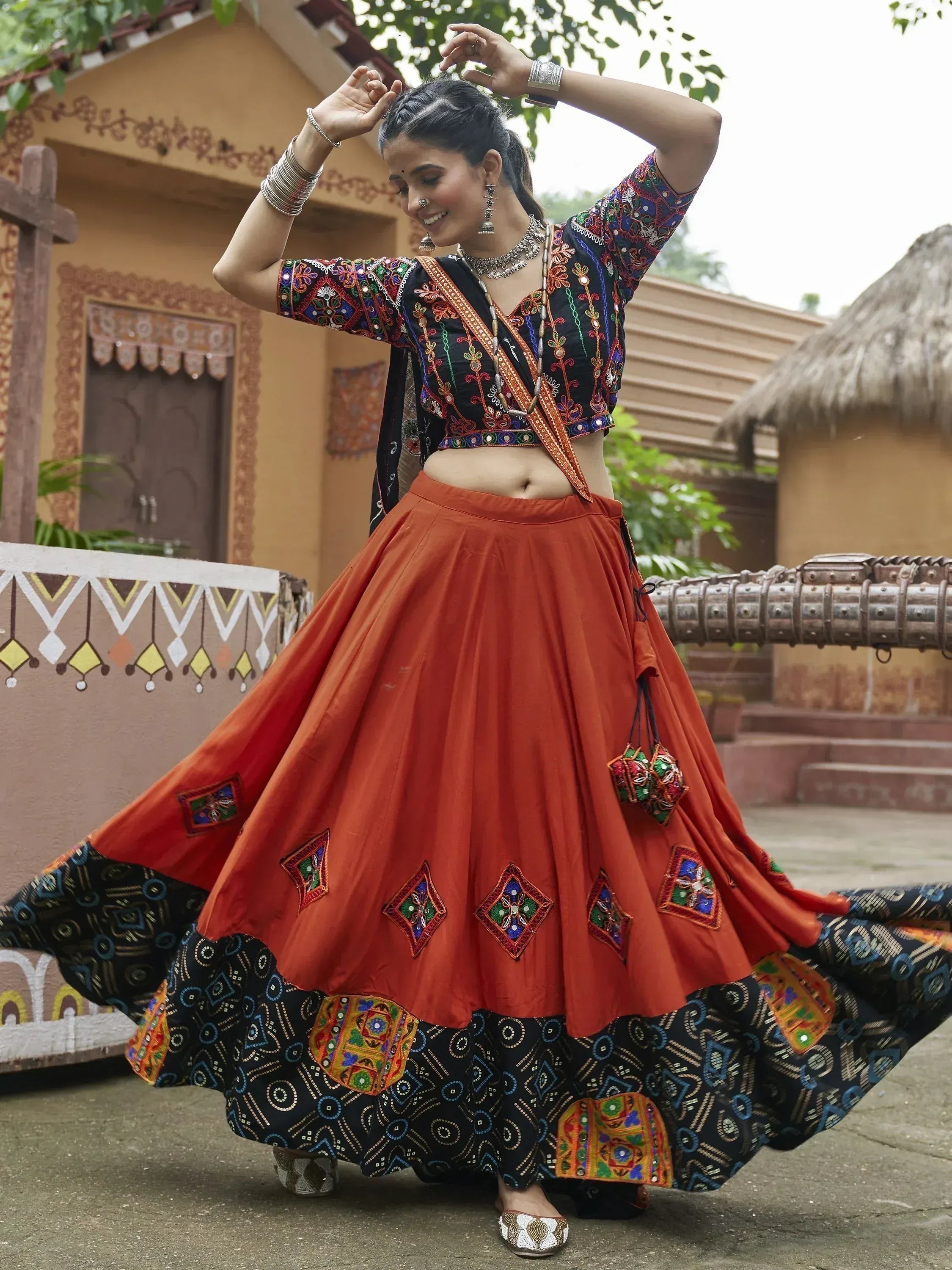 Woman in vibrant Indian ethnic wear, embroidered black top with orange lehenga skirt, traditional jewelry, dancing outdoors