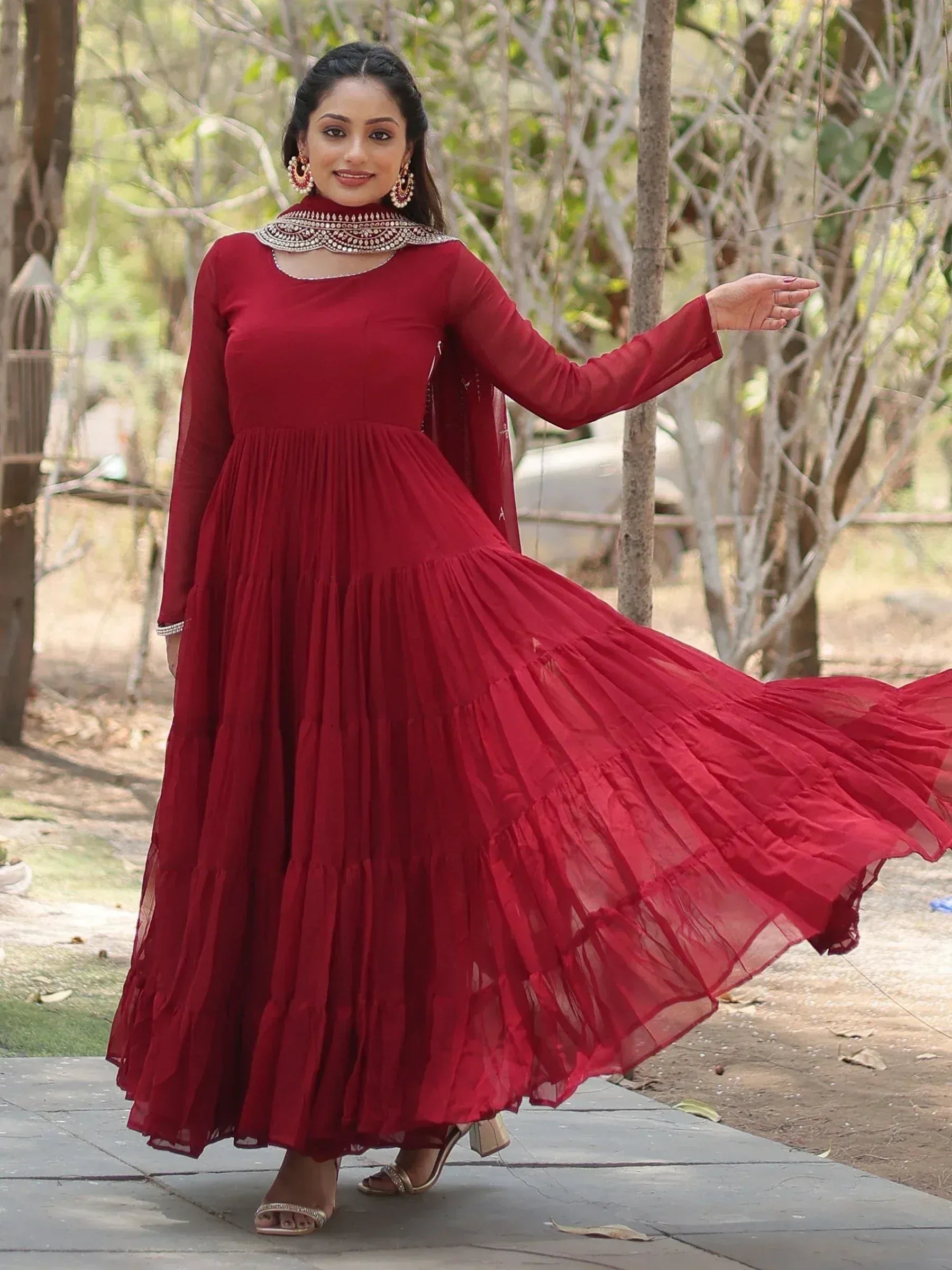 Woman wearing maroon ethnic gown with dupatta outdoors, traditional jewelry, flowy dress