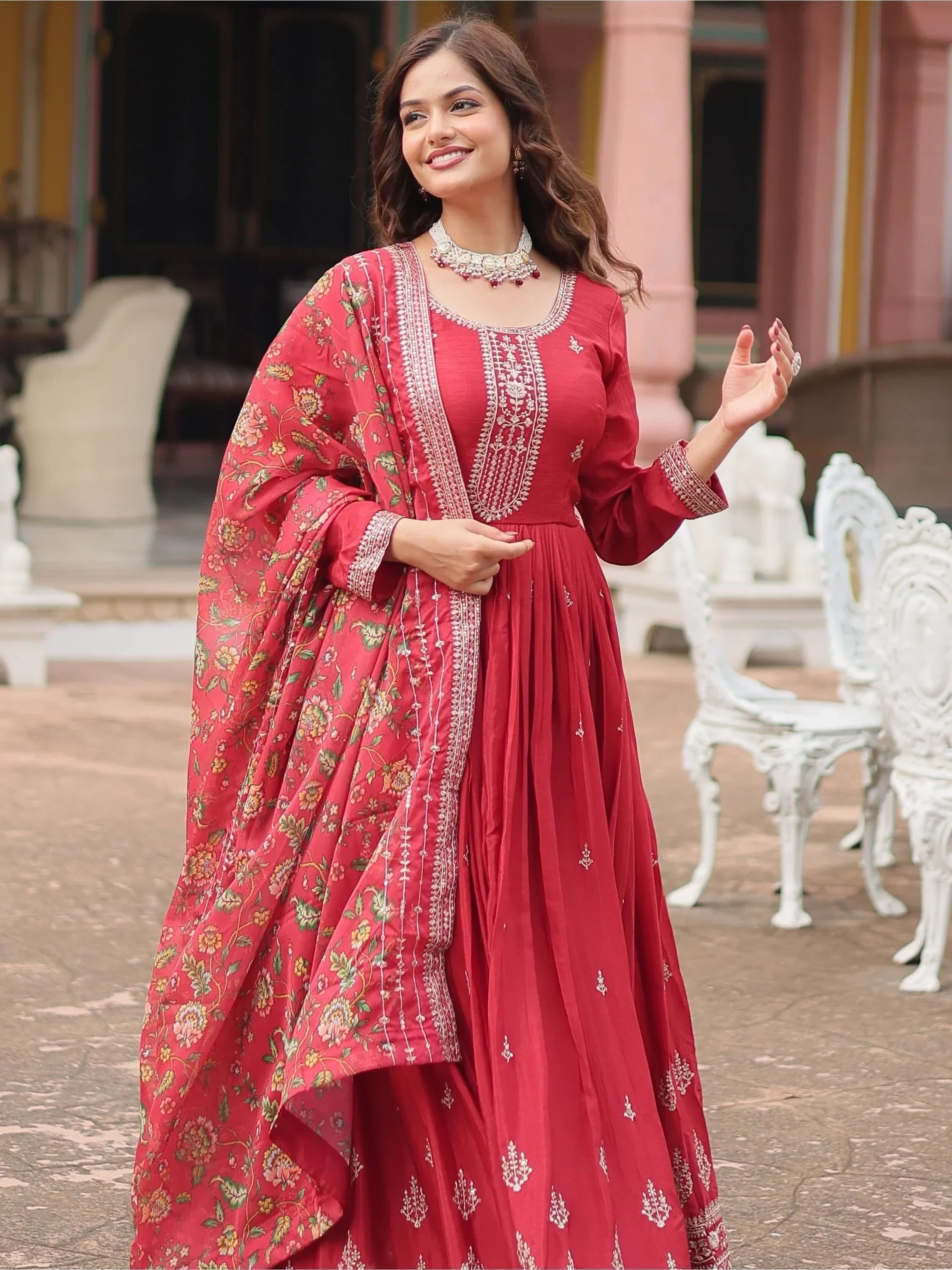 Woman in maroon embroidered ethnic gown with floral dupatta, traditional jewelry outdoors