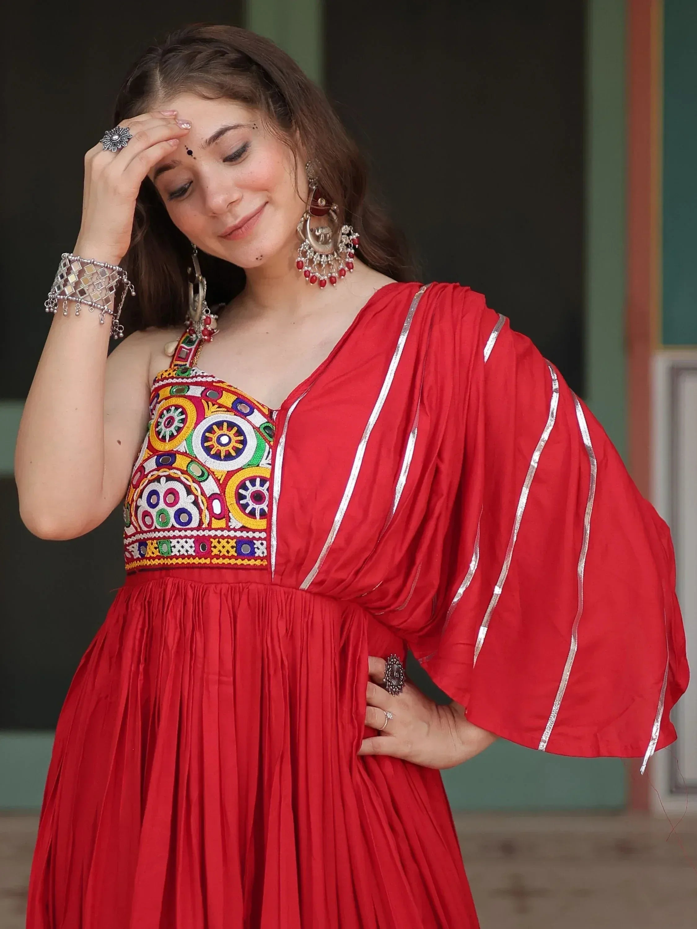 Red ethnic gown with mirror work and silver trim, paired with traditional jewelry for festive occasions