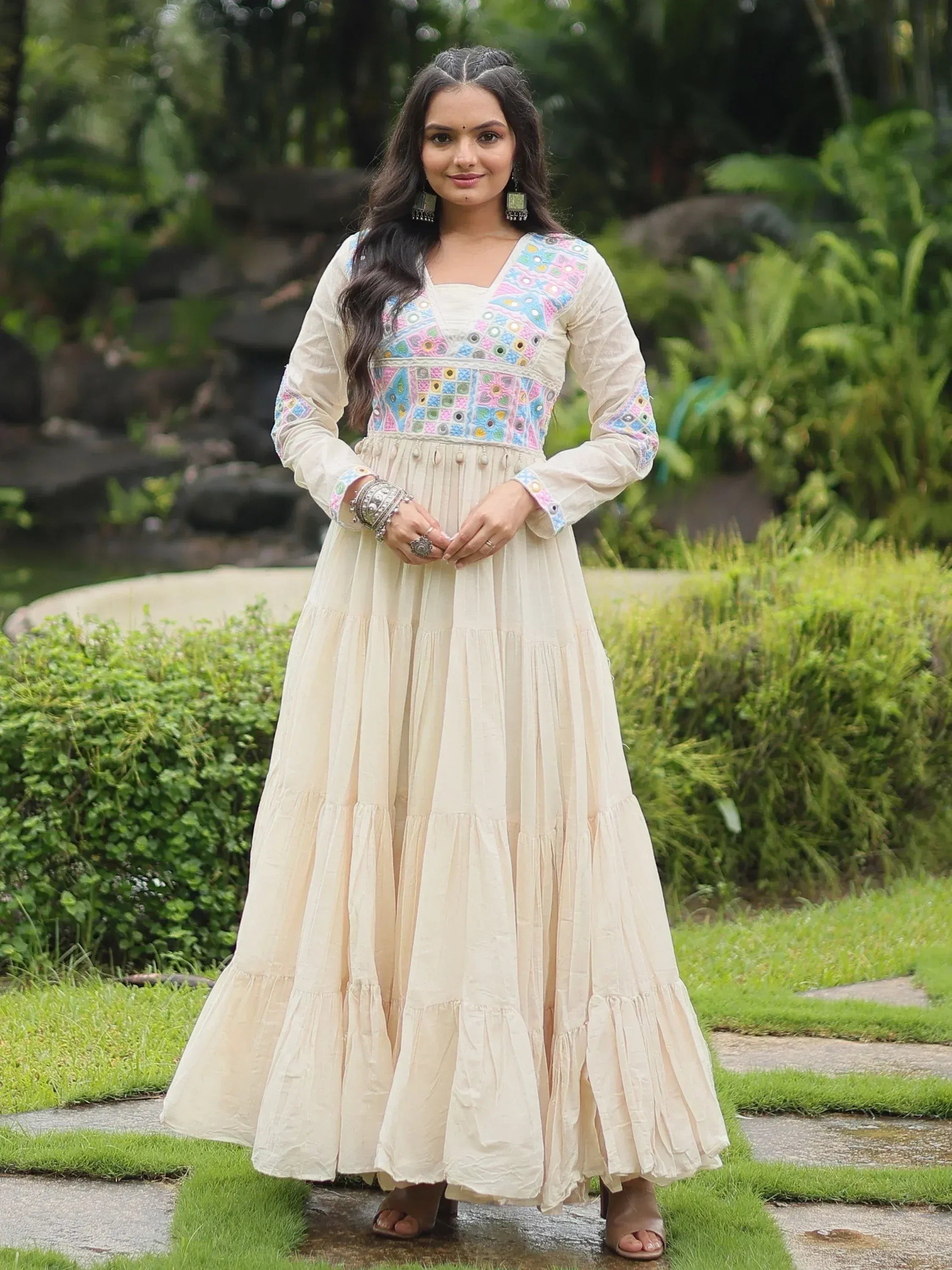 Woman in cream ethnic gown with colorful embroidery, standing outdoors in a garden