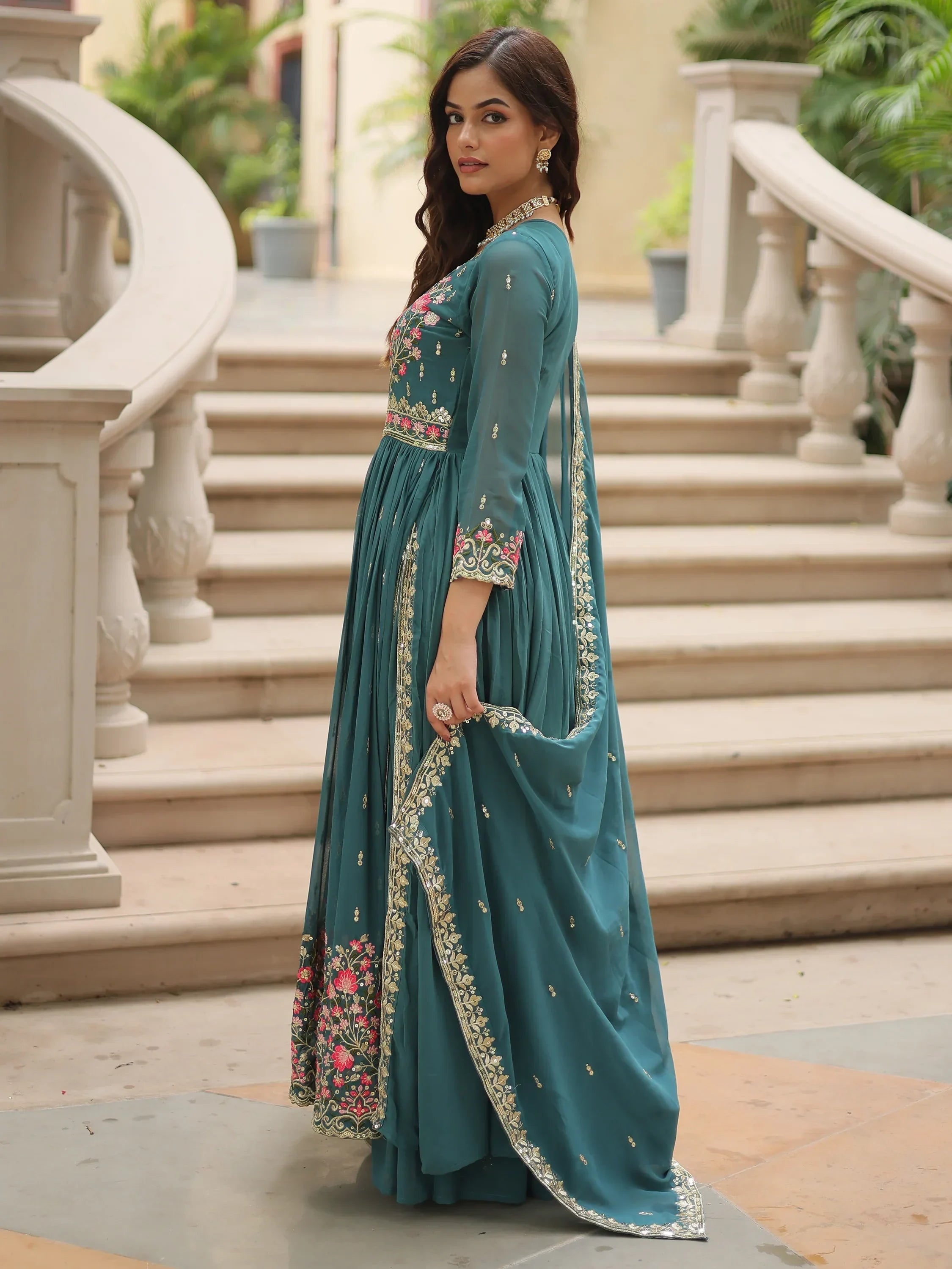 Woman in teal embroidered Anarkali suit with floral details, posing on stone stairs