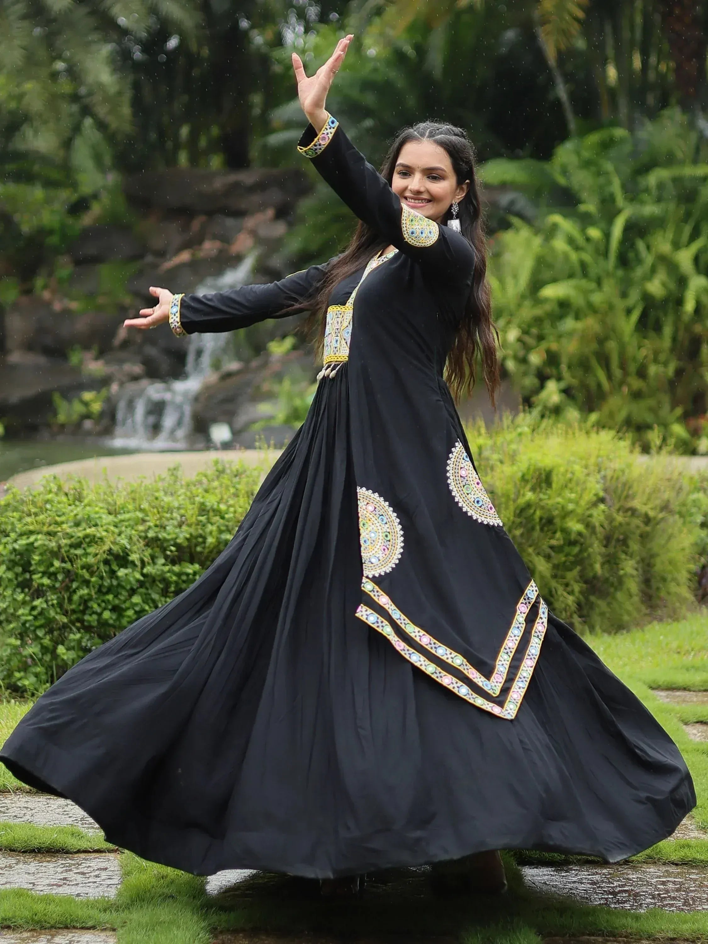Woman twirling in black ethnic gown with colorful embroidery, garden backdrop