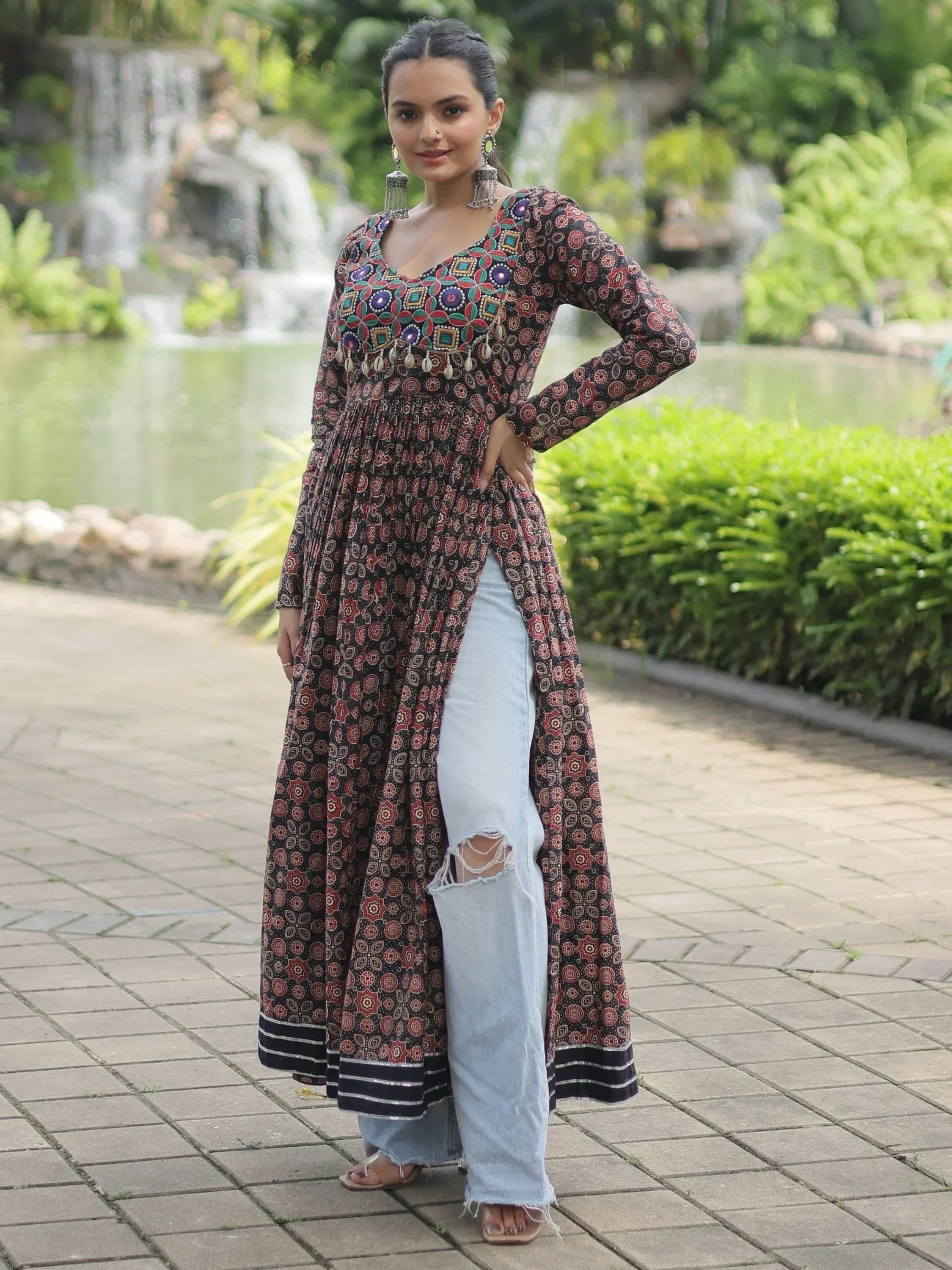 Woman in printed ethnic kurta with mirror work, paired with ripped jeans, standing outdoors near a garden waterfall.