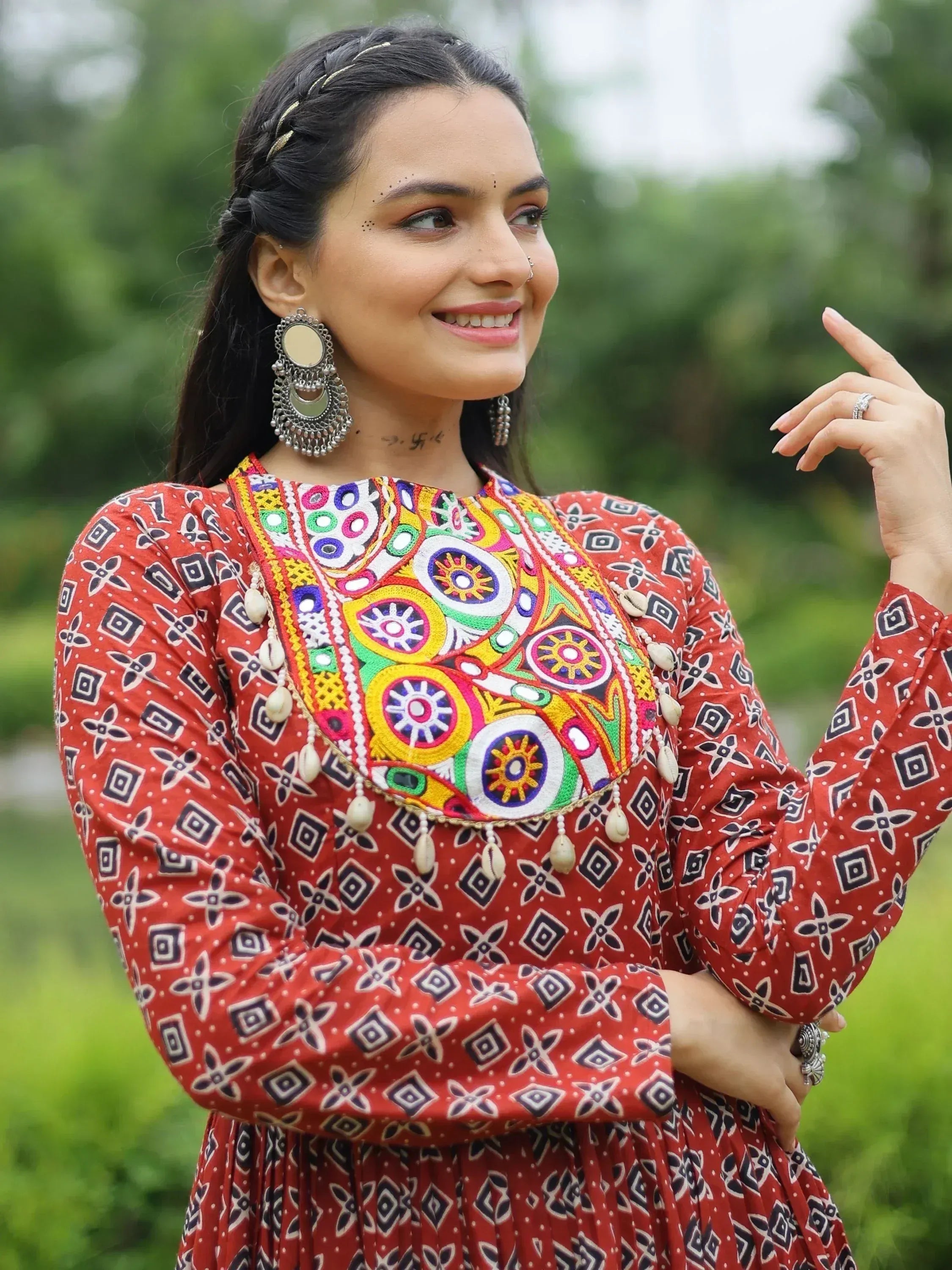 Woman in vibrant red ethnic dress with colorful mirror embroidery and traditional jewelry, styled for festive occasions.