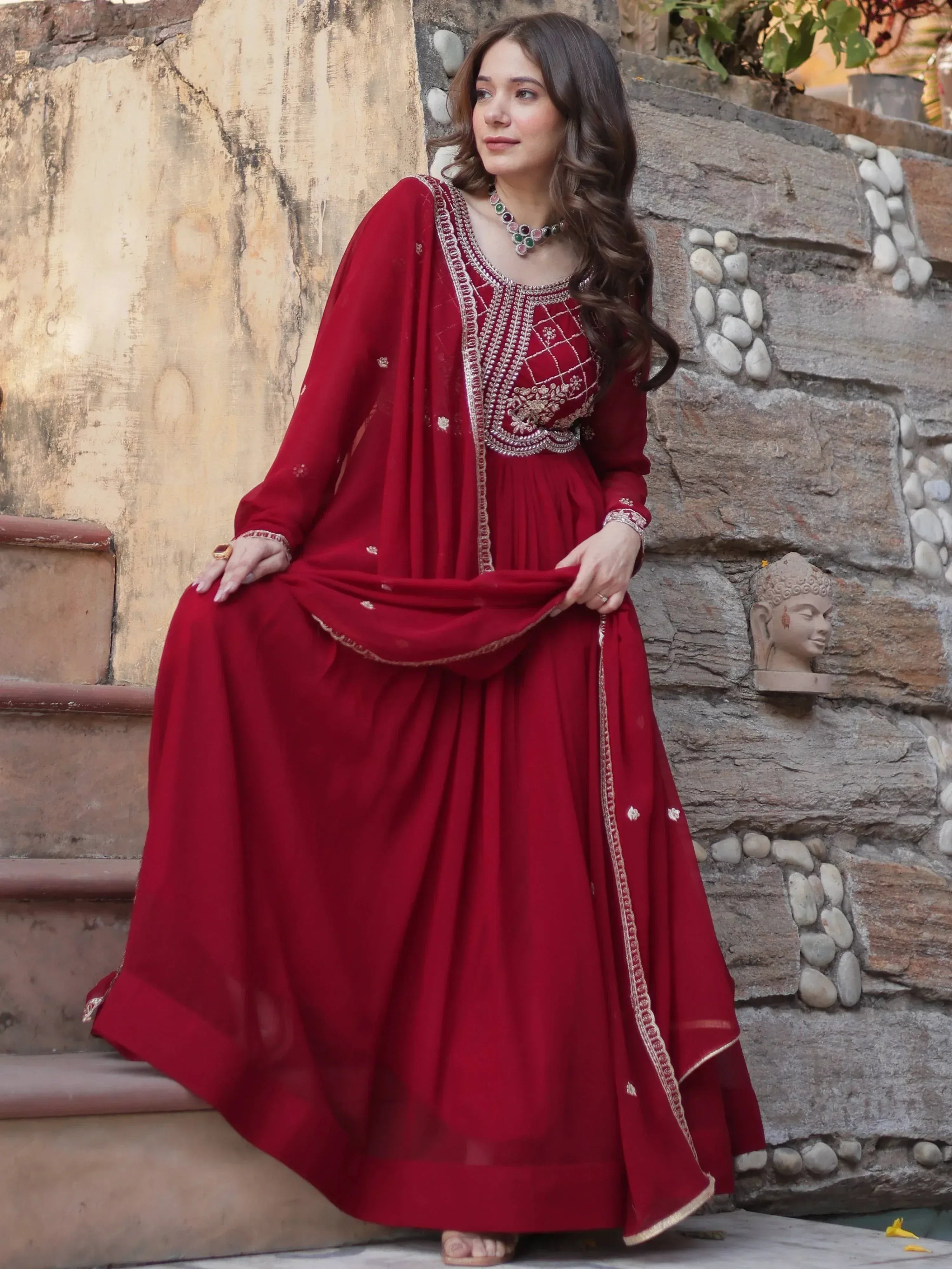 Woman in red embroidered Indian anarkali suit with dupatta, standing by stone wall