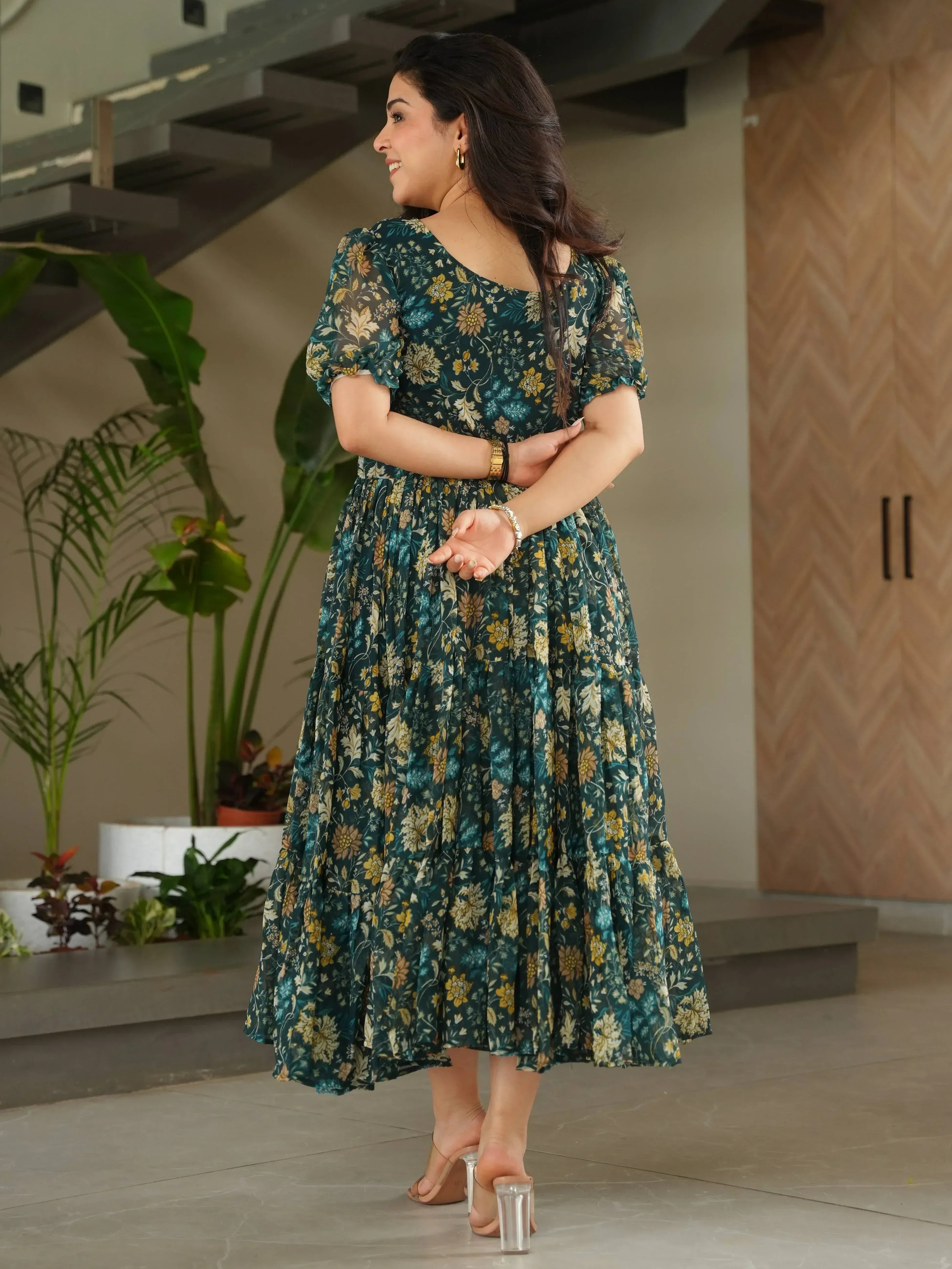 Woman in green floral ethnic gown with puff sleeves, standing indoors near plants and modern staircase