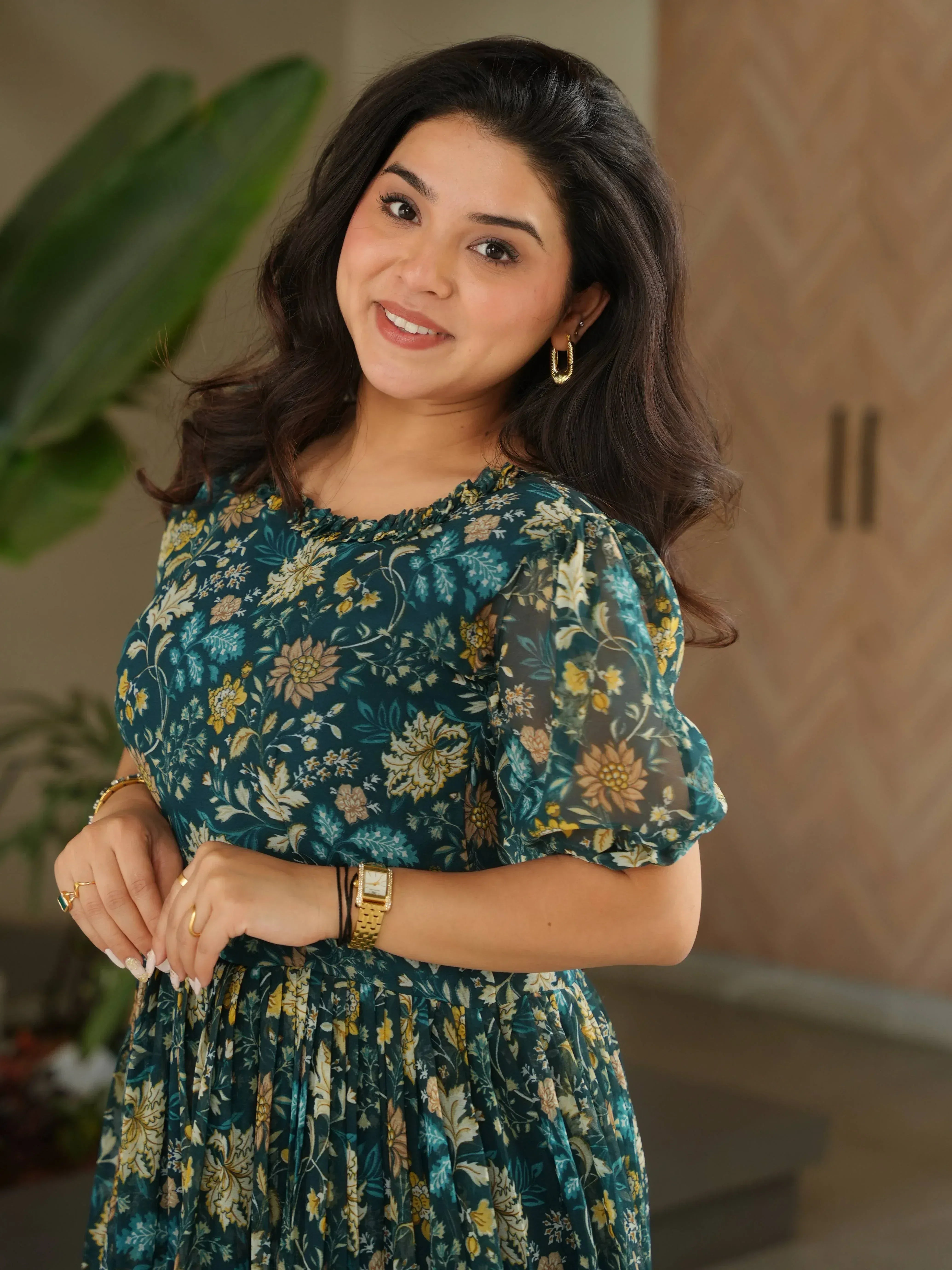 Woman in green floral ethnic gown with puff sleeves, gold jewelry, smiling indoors