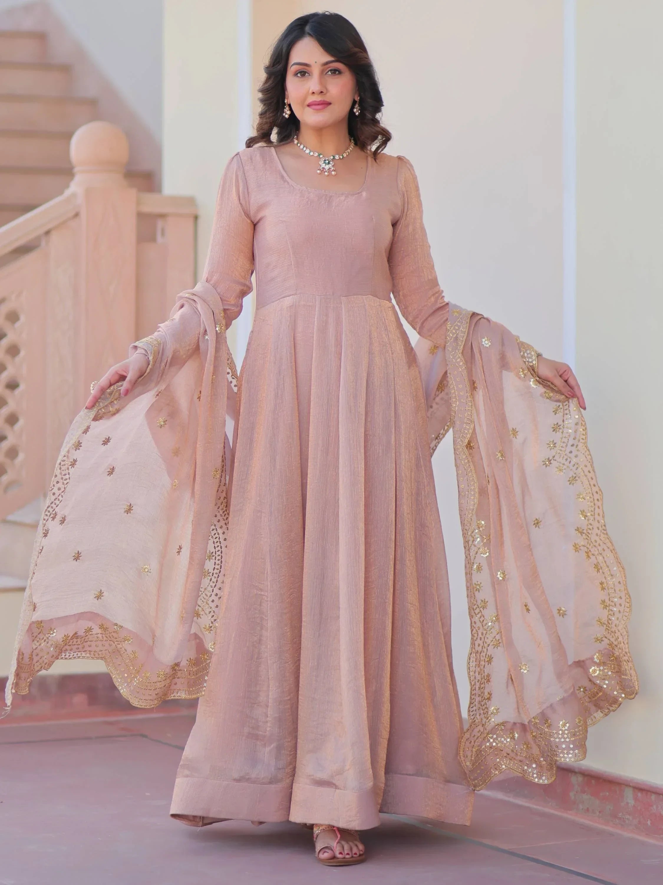 Woman wearing a pastel pink ethnic gown with embroidered dupatta, standing outdoors.