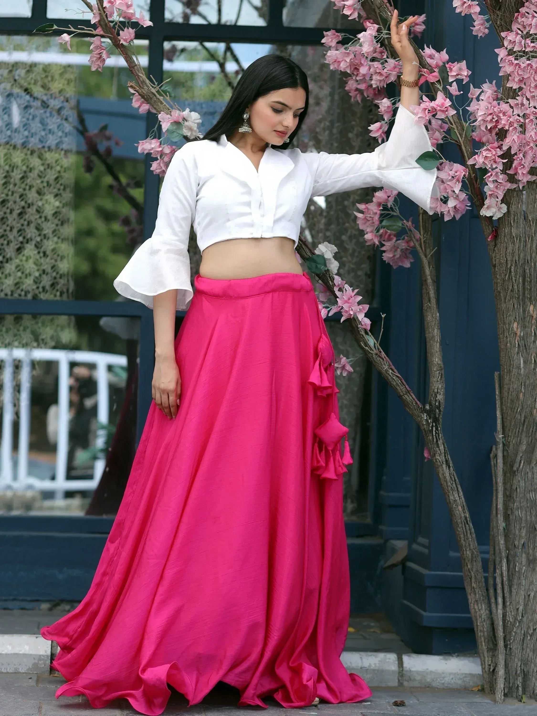 Woman in white crop top and vibrant pink lehenga skirt with ruffles, standing near pink flowers