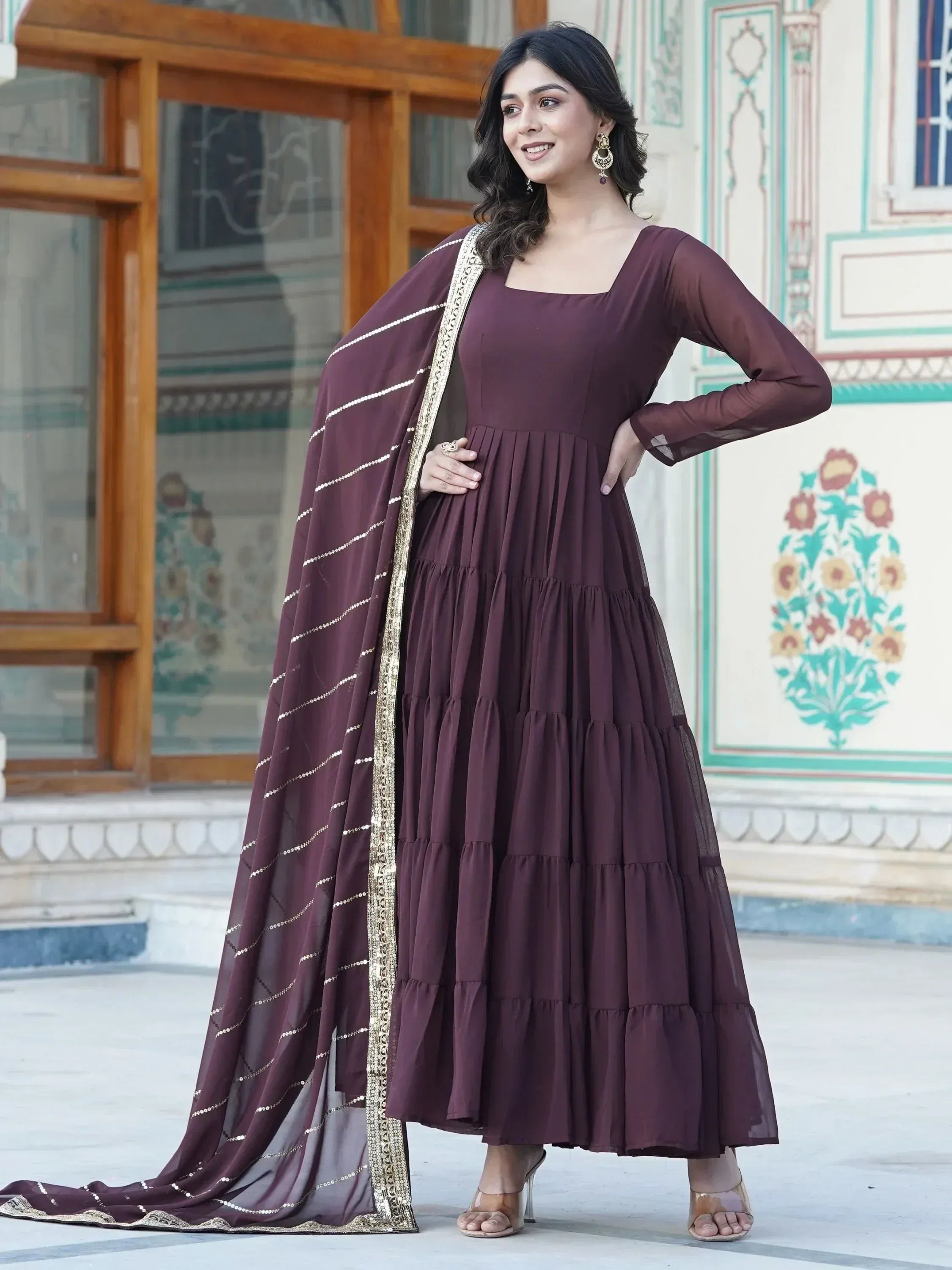 Woman in brown tiered ethnic Anarkali gown with sheer dupatta and gold trim, standing outdoors