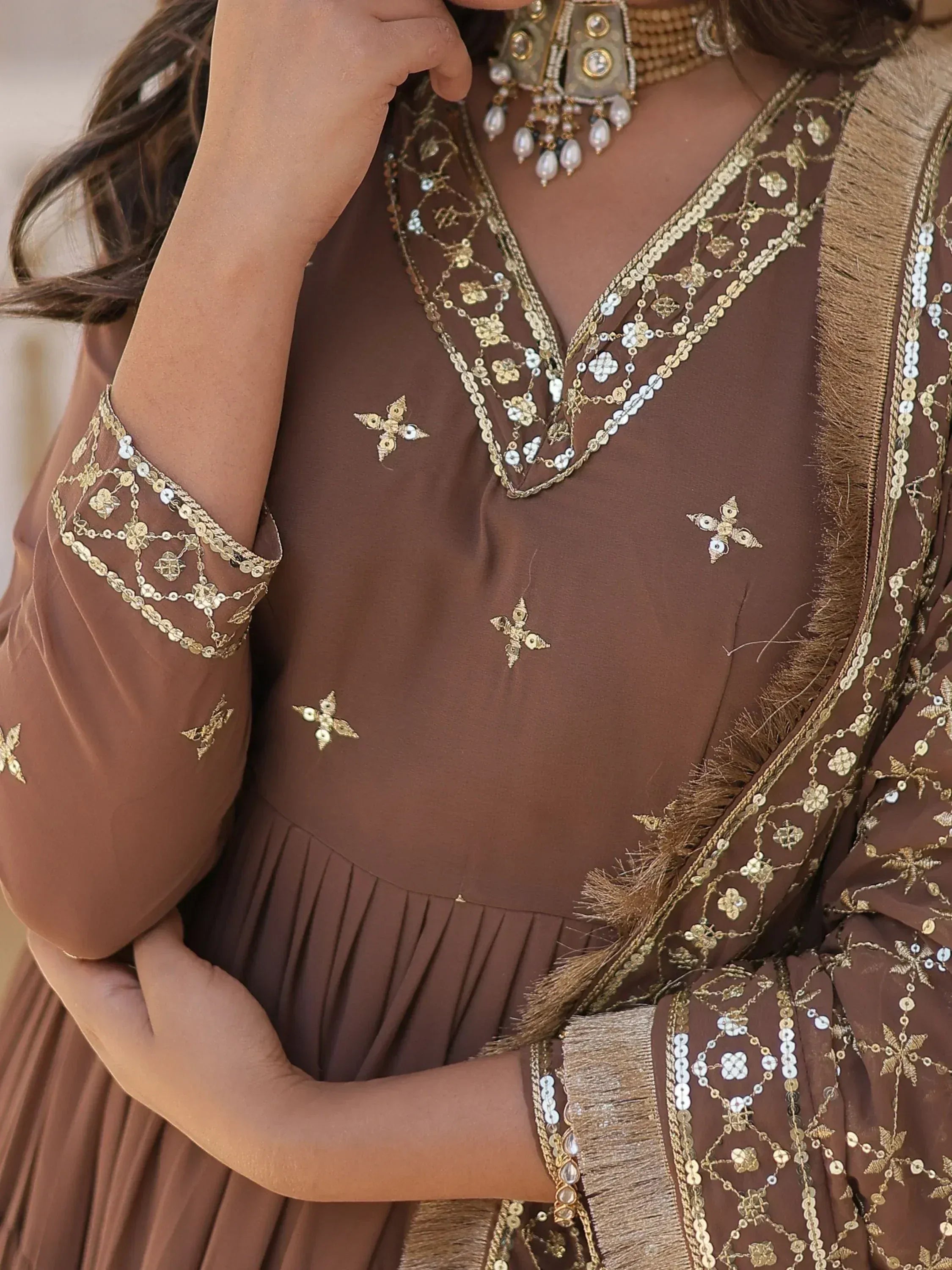 Brown ethnic gown with gold mirror work, sequins, and fringe detailing, paired with traditional jewelry.