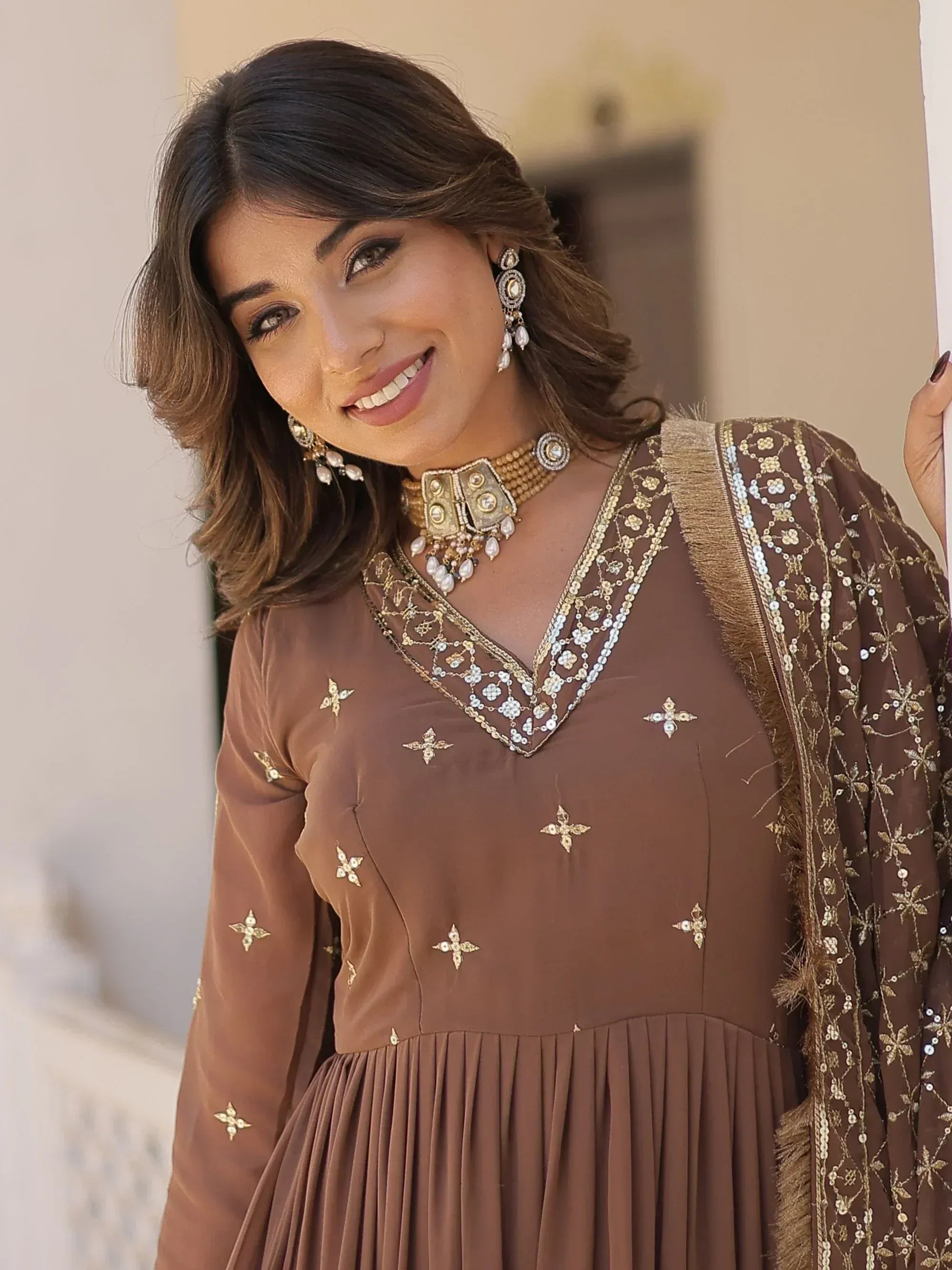 Brown embroidered ethnic gown with gold detailing, paired with traditional jewelry for festive occasions.