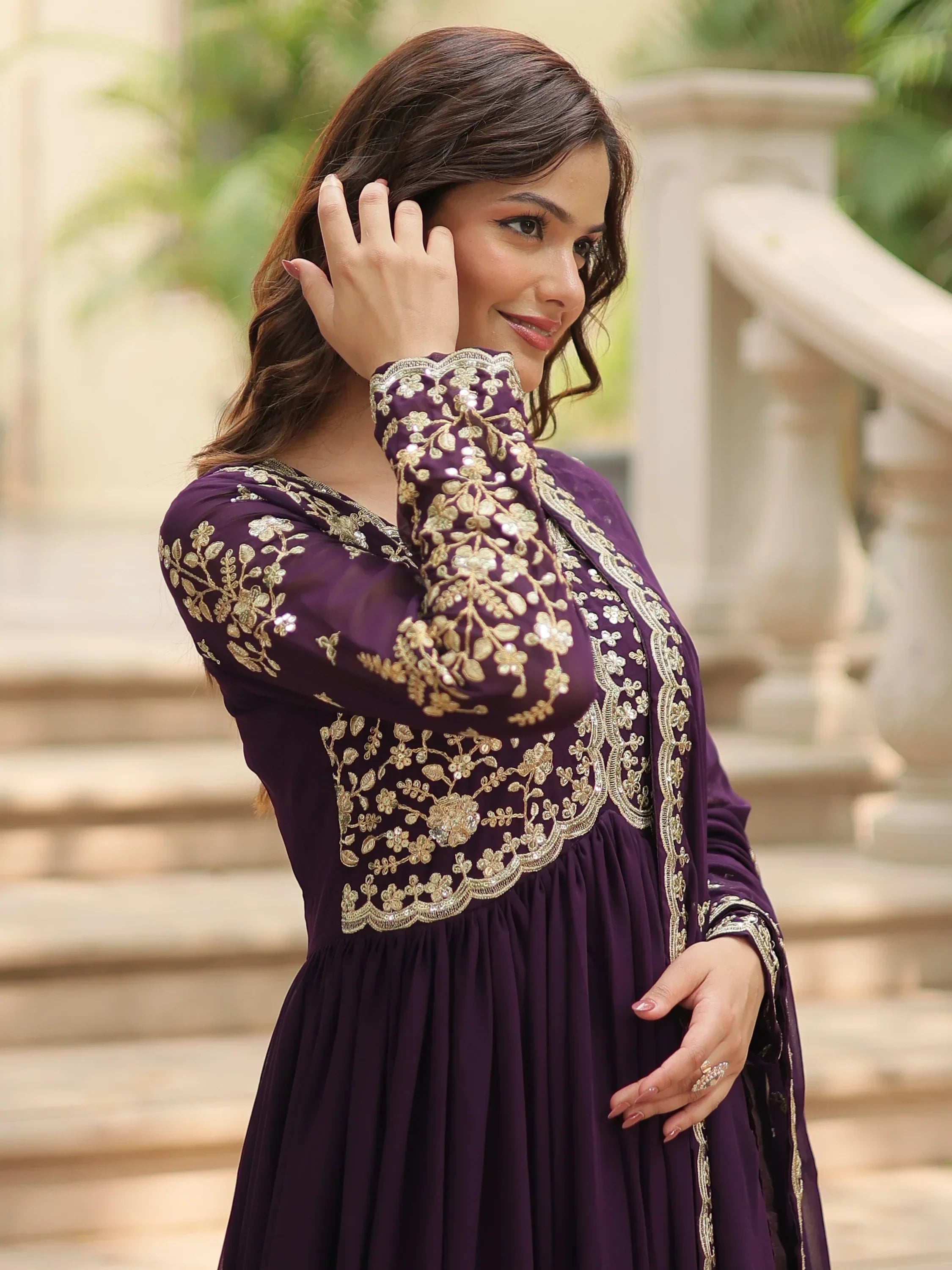 Woman in a purple ethnic gown with gold embroidery, standing outdoors by stone steps