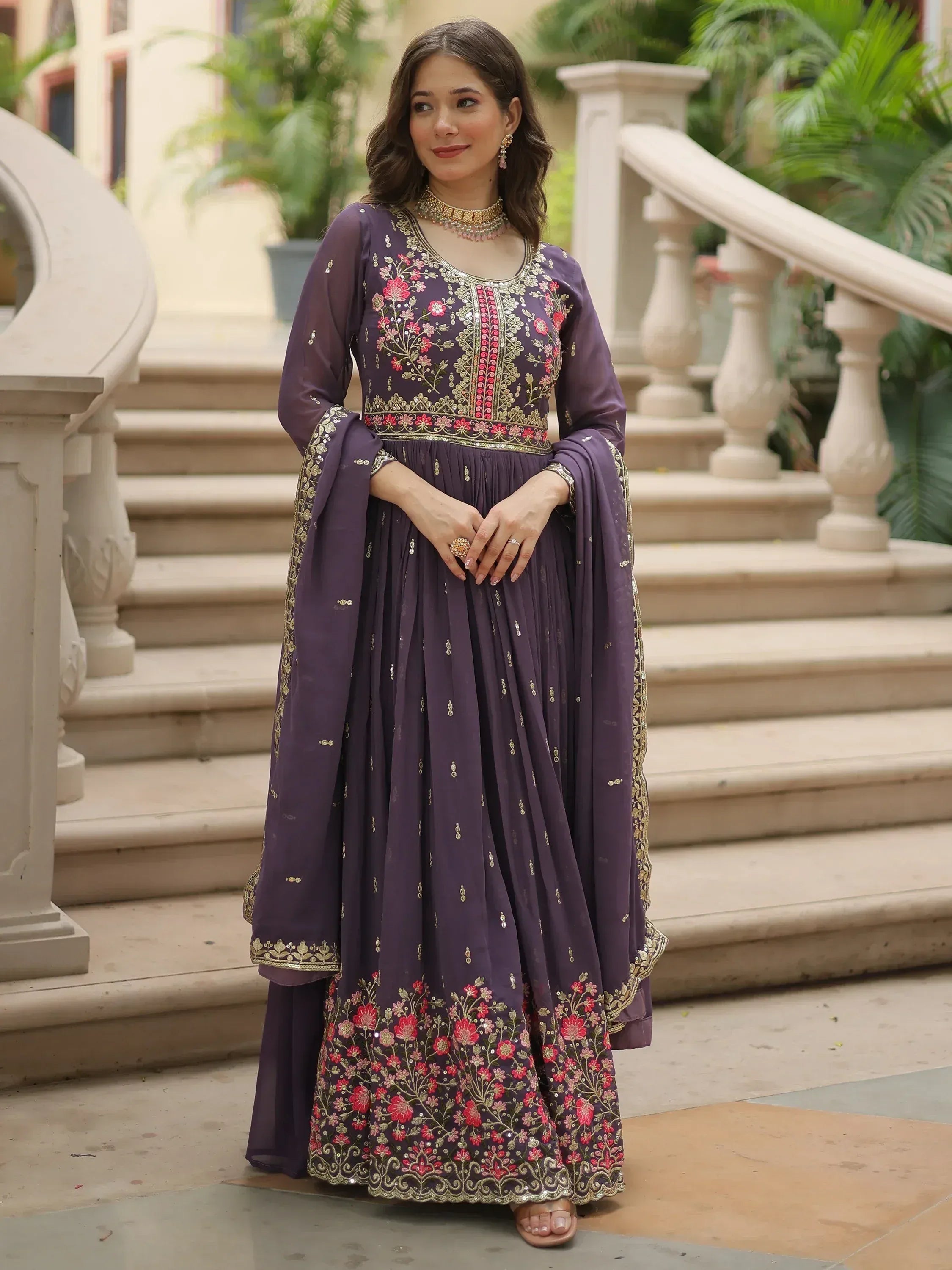 Woman wearing a purple embroidered ethnic gown with floral detailing, standing outdoors.