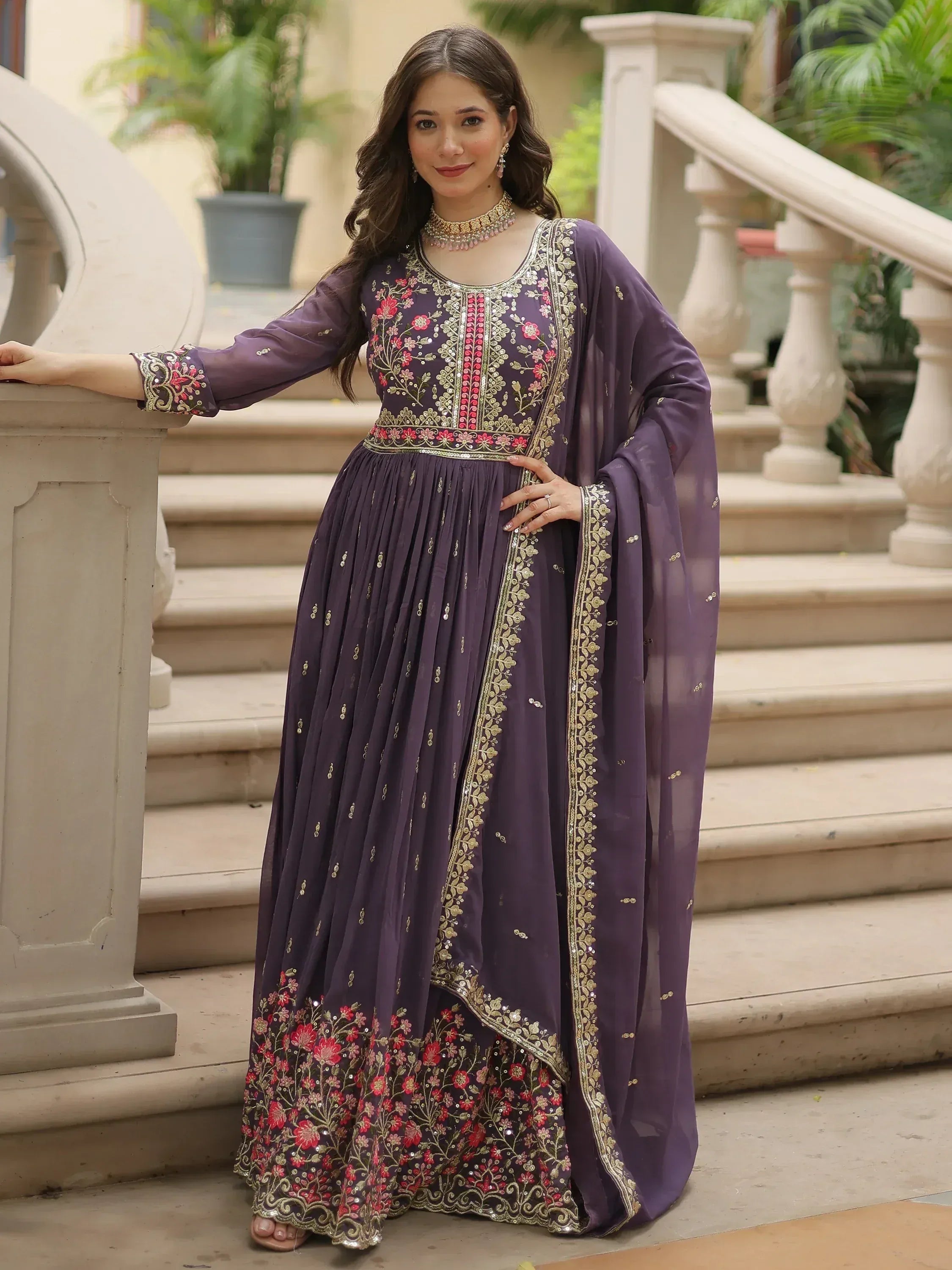 Woman in purple embroidered Anarkali gown with floral designs, standing on outdoor stairs