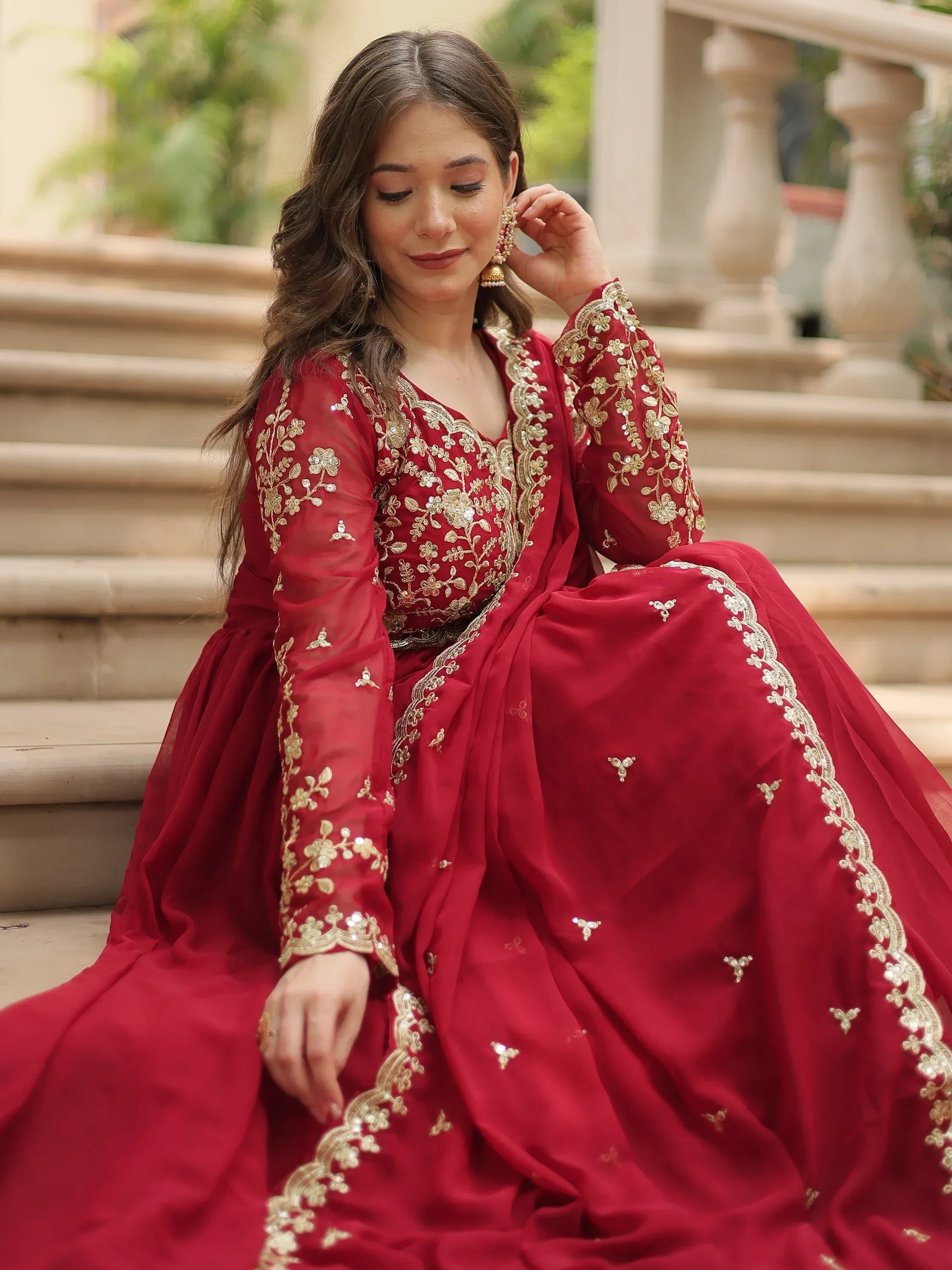 Woman in red embroidered lehenga choli with gold floral details, sitting on steps
