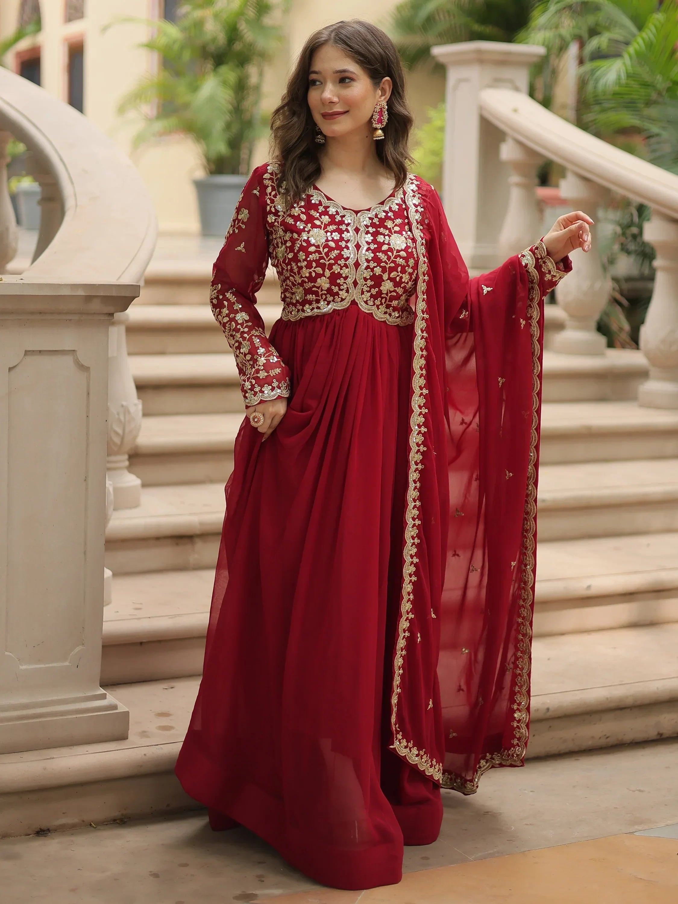 Woman in elegant red embroidered Anarkali suit with dupatta, standing on outdoor steps