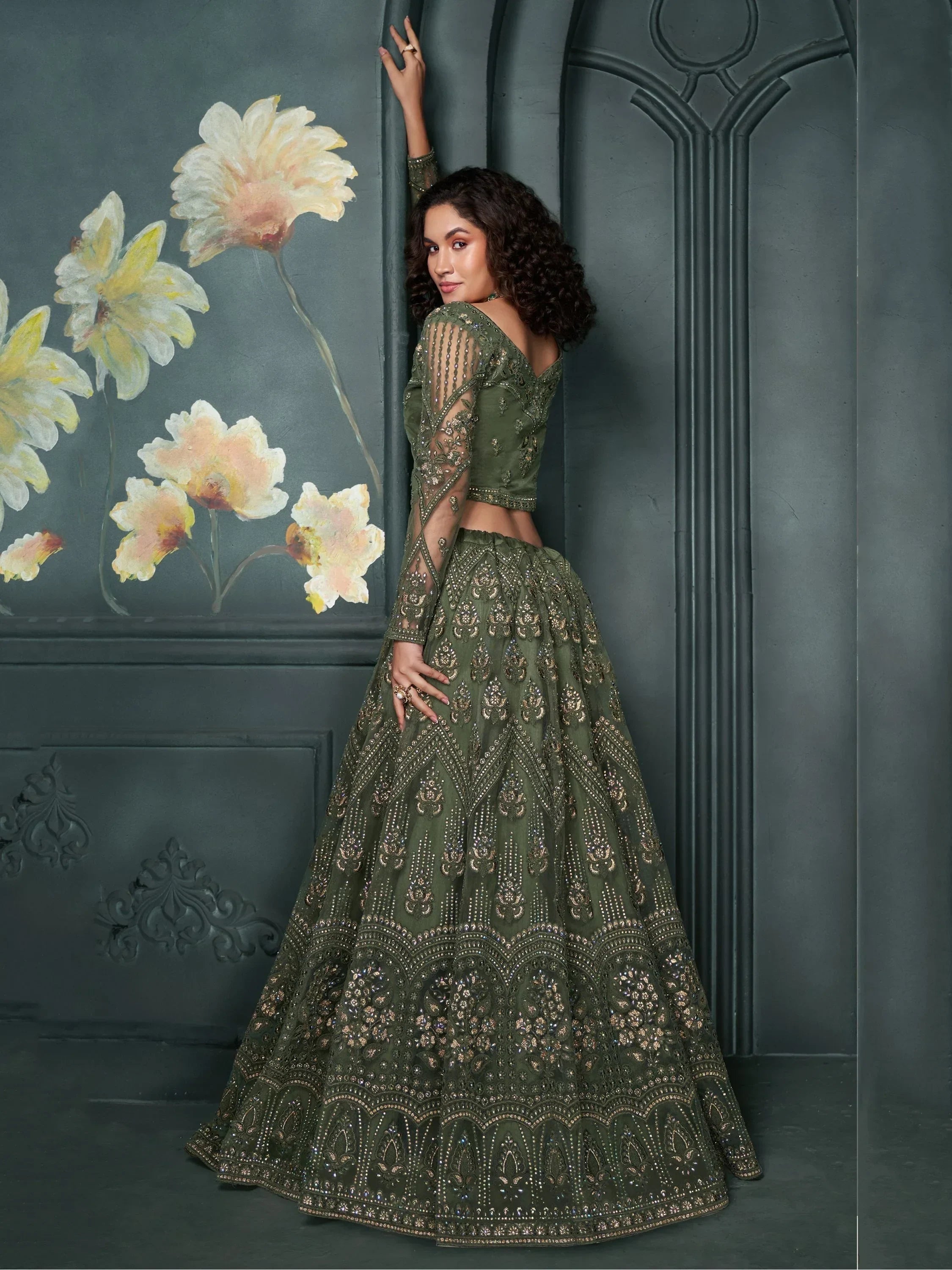 Olive green designer lehenga choli with heavy gold embroidery, sheer sleeves, and floral wall backdrop