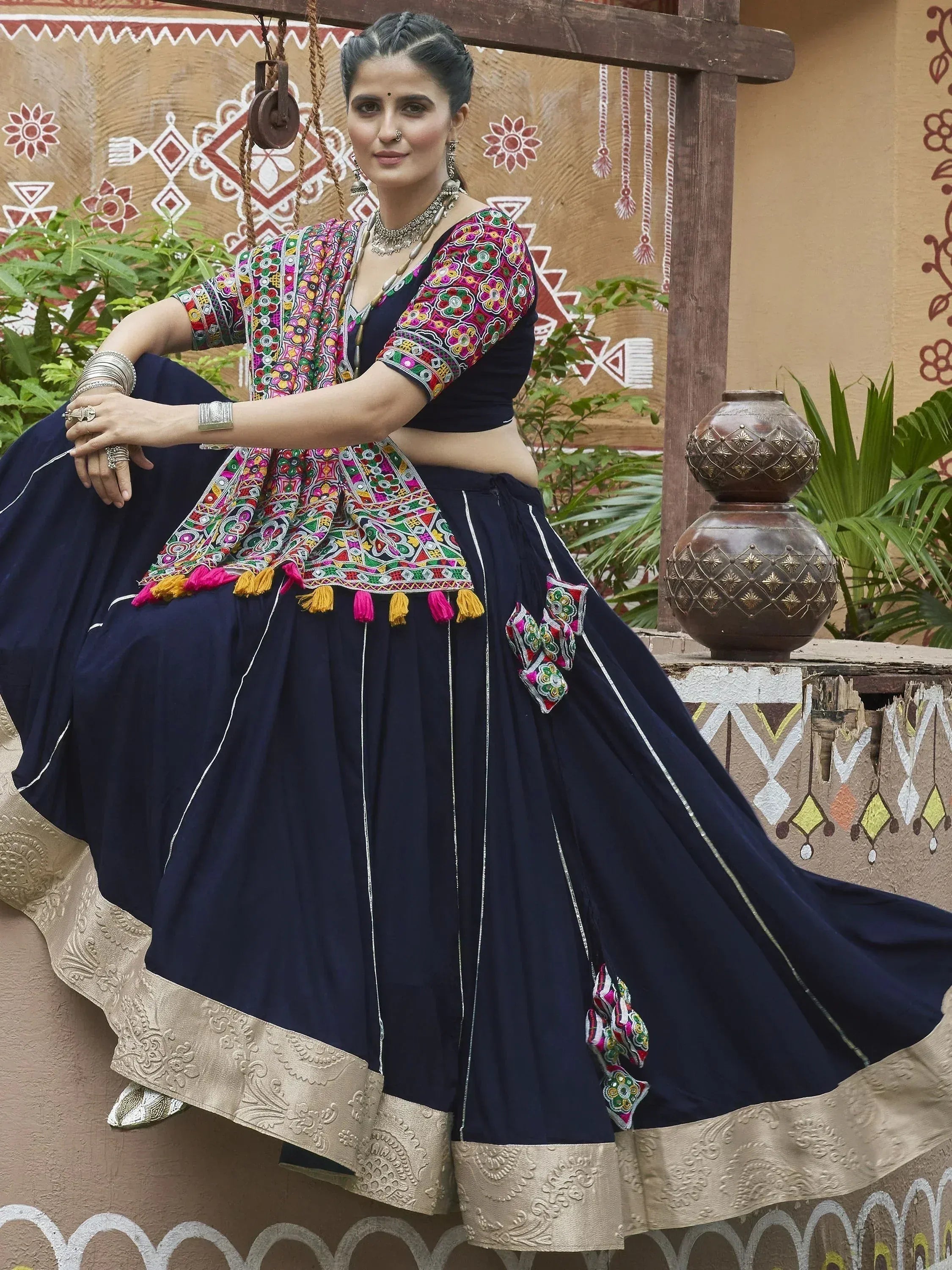 Woman in navy blue embroidered lehenga choli with colorful mirror work, tassels, and gold border, styled with traditional jewelry.